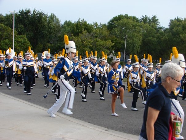 A drum major in a white uniform and tall feathered hat leads a marching band in a parade.