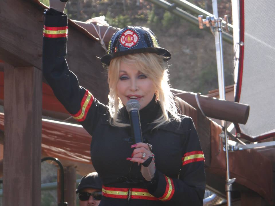Blonde woman in firefighter costume holding a microphone on an outdoor stage.