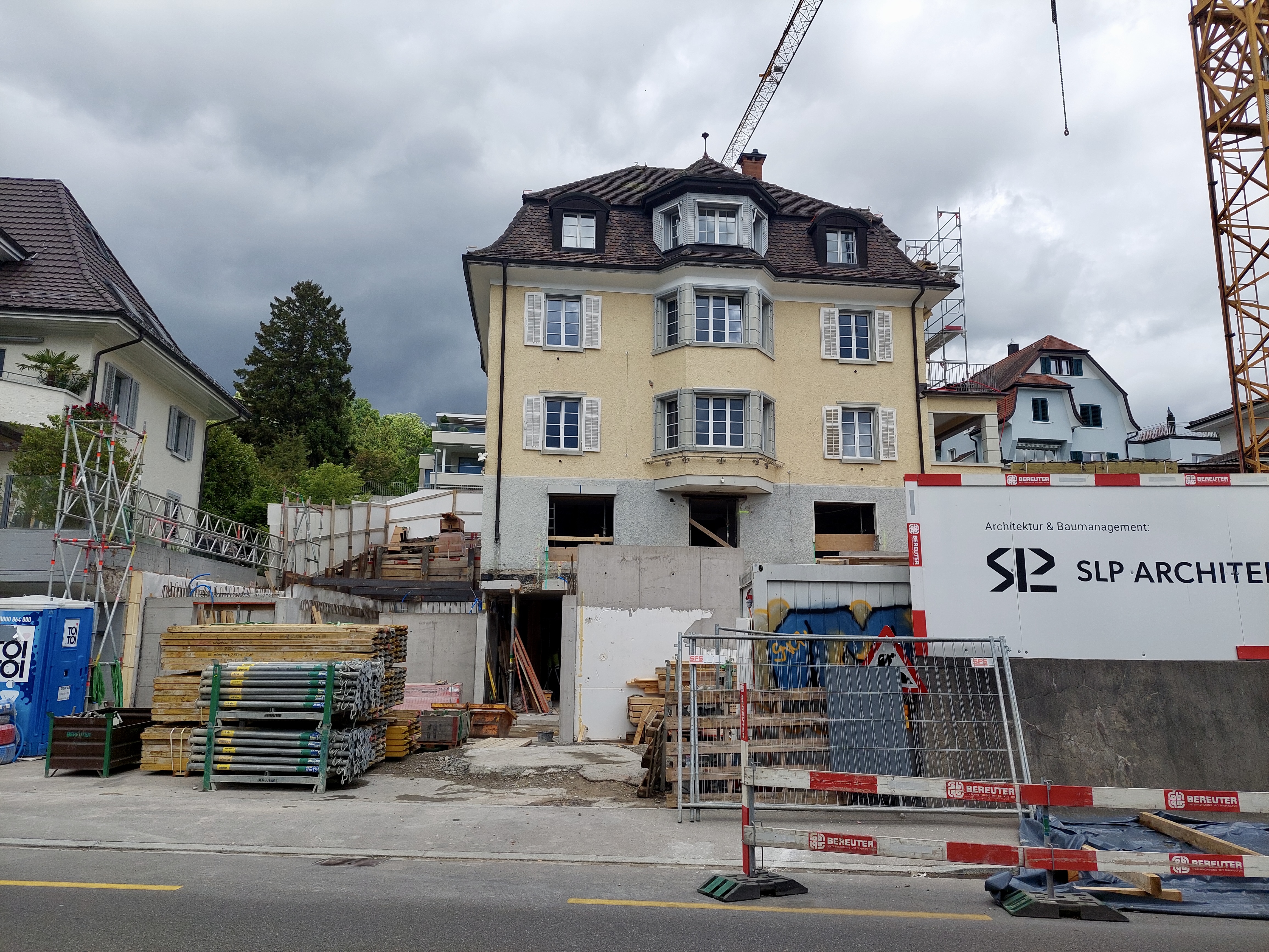 Multi-story building surrounded by scaffolding, lumber, construction fencing, and a tower crane.