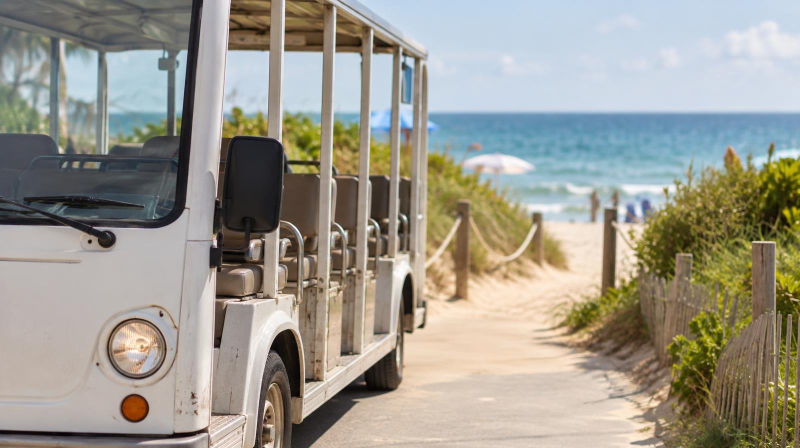 Small open-air trolley bus parked beside a sandy beach path with ocean beyond.