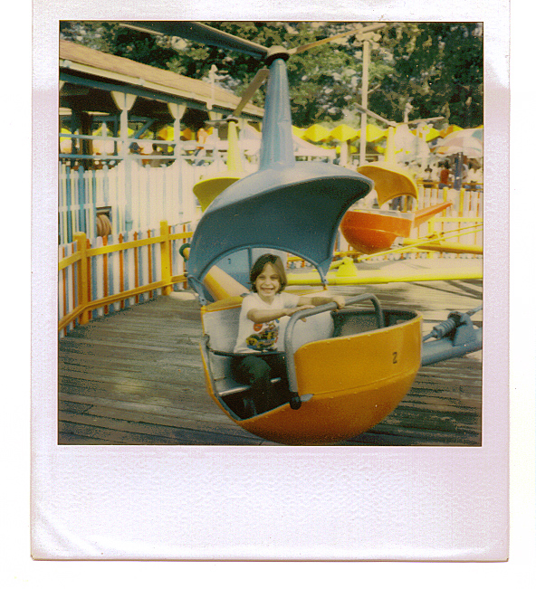 A child sitting in a yellow and blue helicopter-shaped kiddie ride at an amusement park.