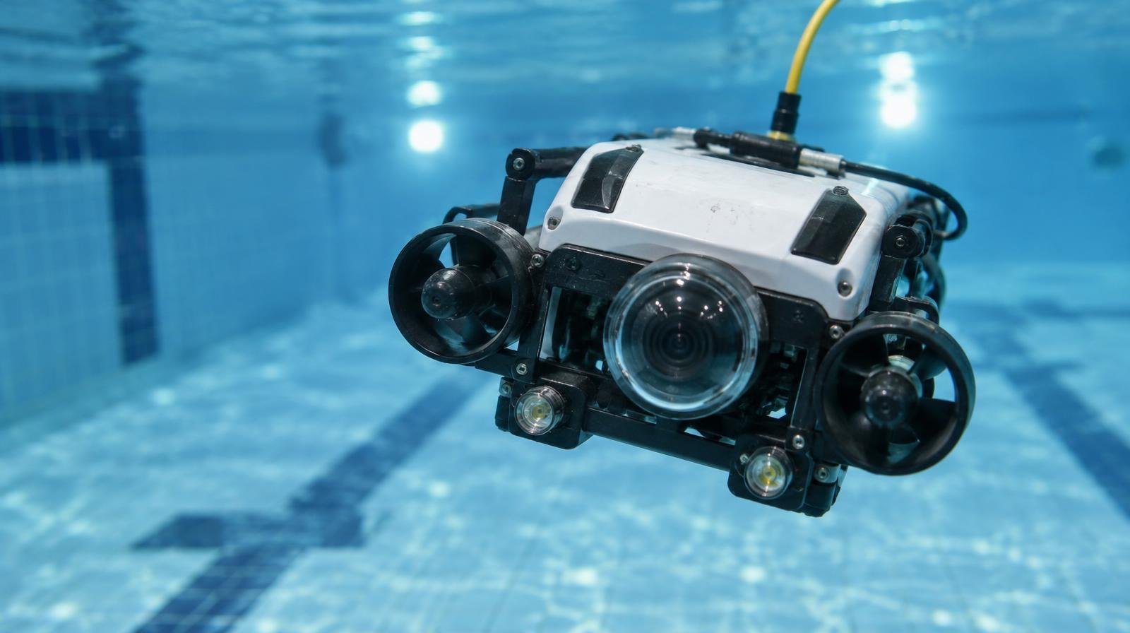 A small underwater robot with thrusters and a camera submerged in clear blue water.