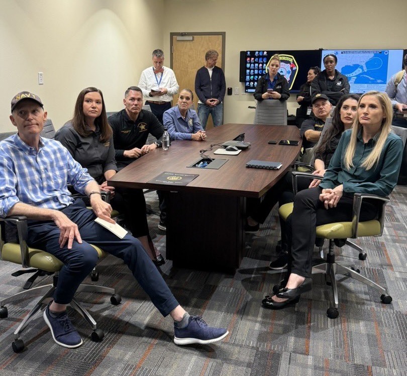 Officials seated around a conference table inside an emergency operations center, map displayed on screen behind them.
