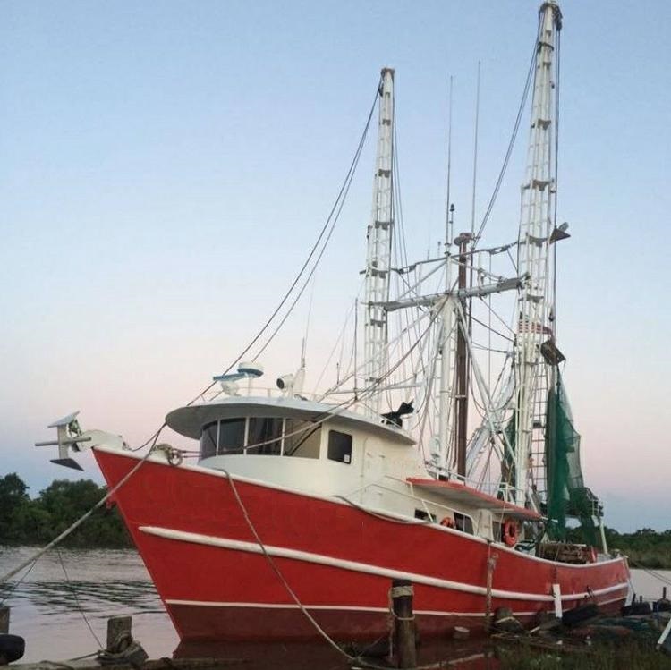 Red-hulled shrimp trawler with tall outrigger booms and green nets moored at a dock.