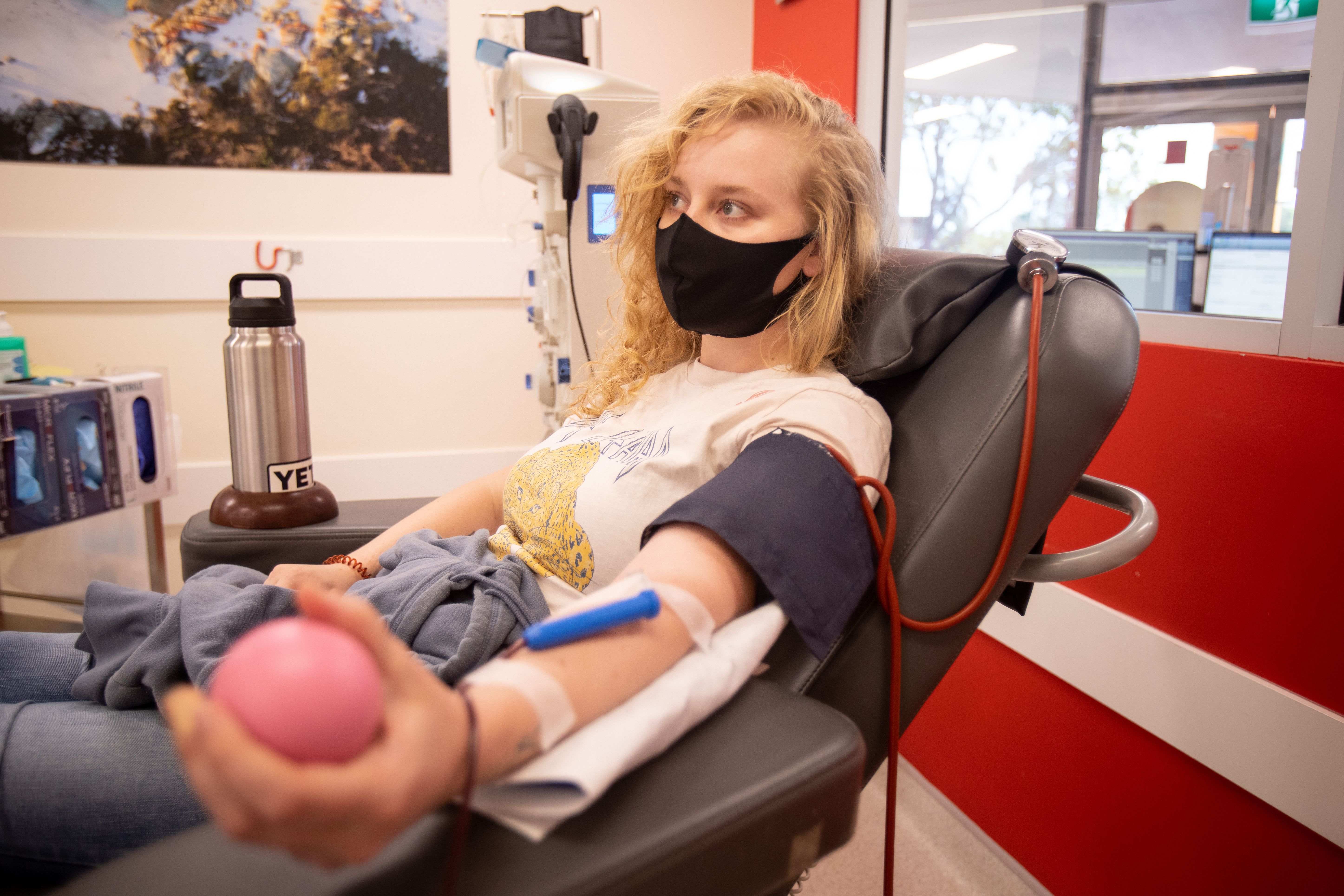 Masked person seated in a donation chair squeezes a pink stress ball with an IV line in their arm.