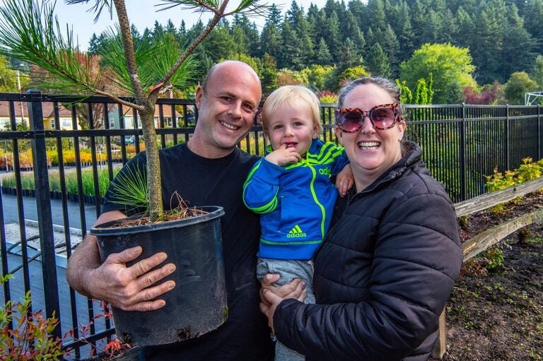 A man holds a potted young tree while a woman and toddler stand beside him outdoors near a metal fence.