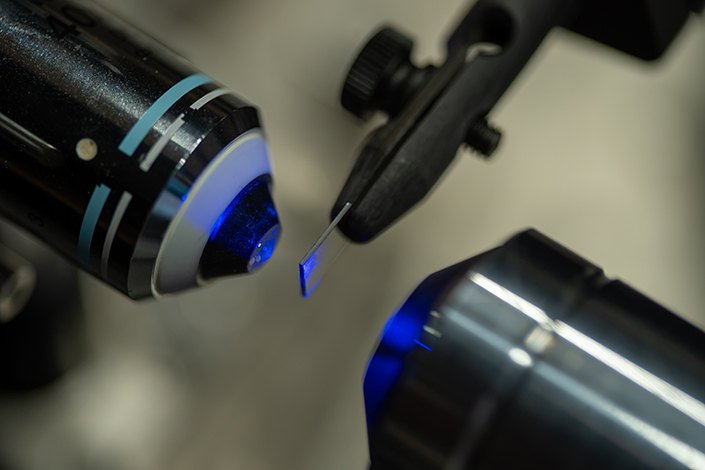 A tiny metallic strip clamped between two probe tips, illuminated by blue light from nearby optical equipment.