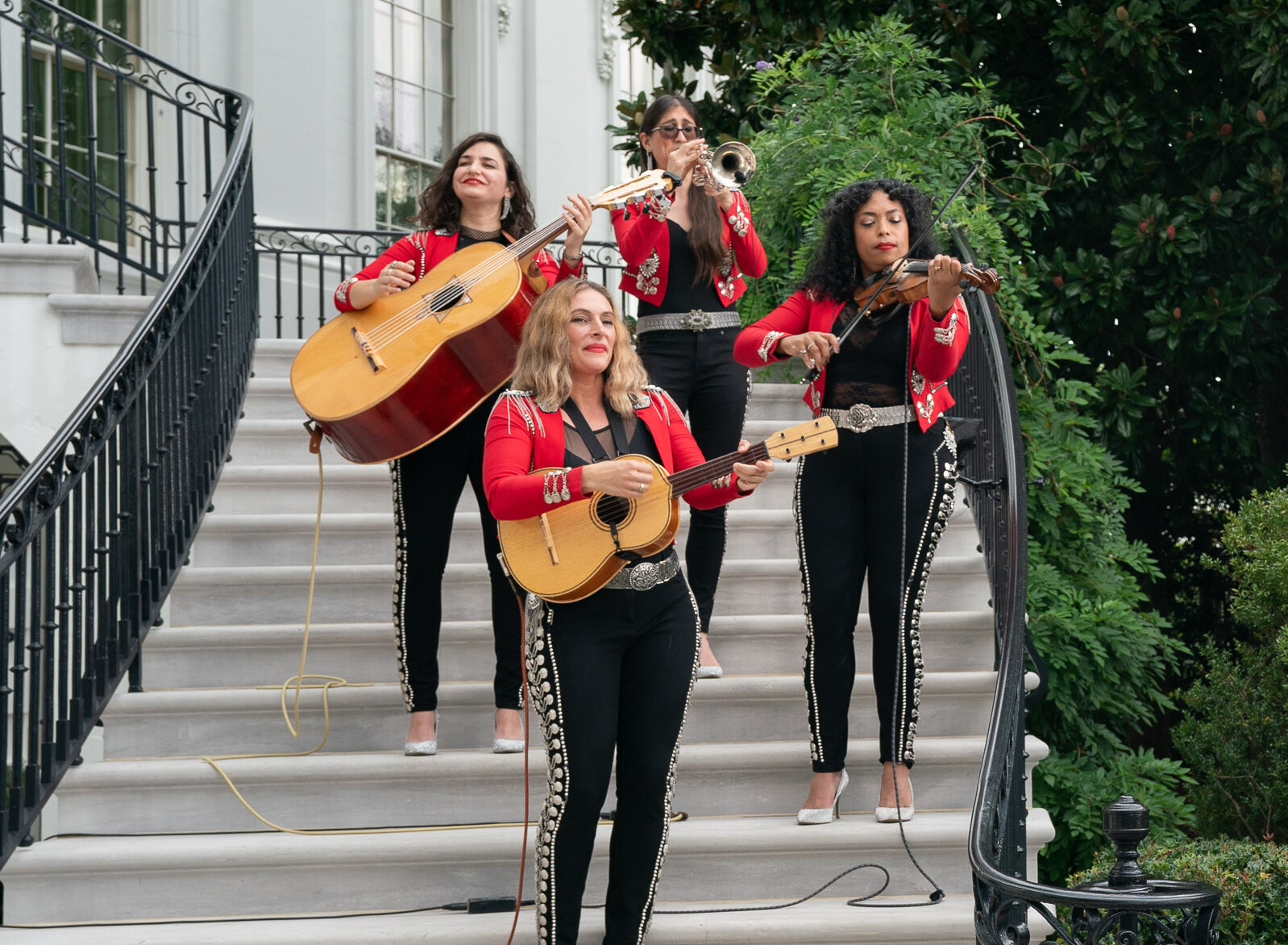 Four women in red mariachi jackets perform with string instruments on outdoor steps.