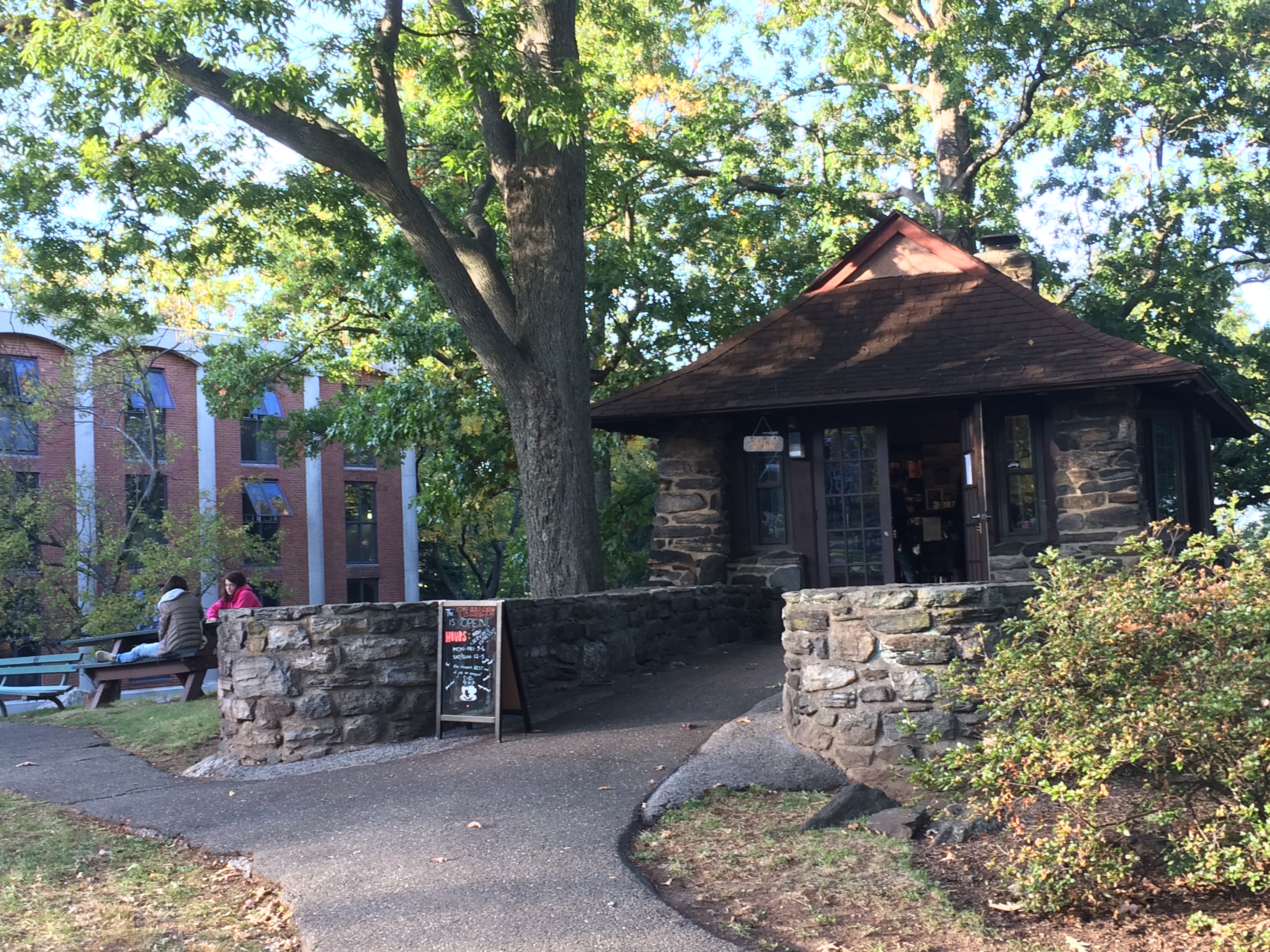 Small stone building on a tree-lined campus path with a brick building in the background.