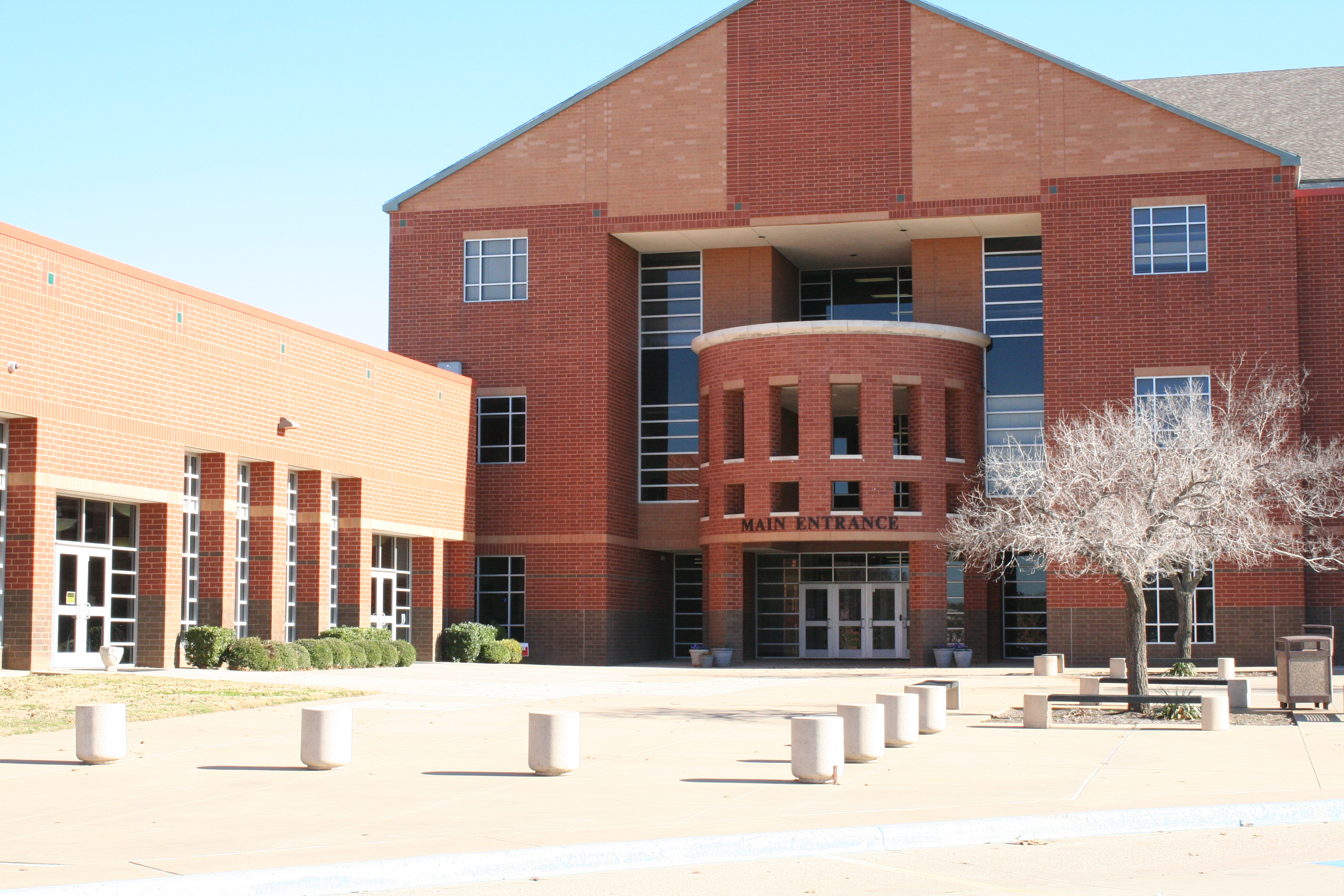 Exterior of a red-brick high school building with a curved main entrance and bollards.