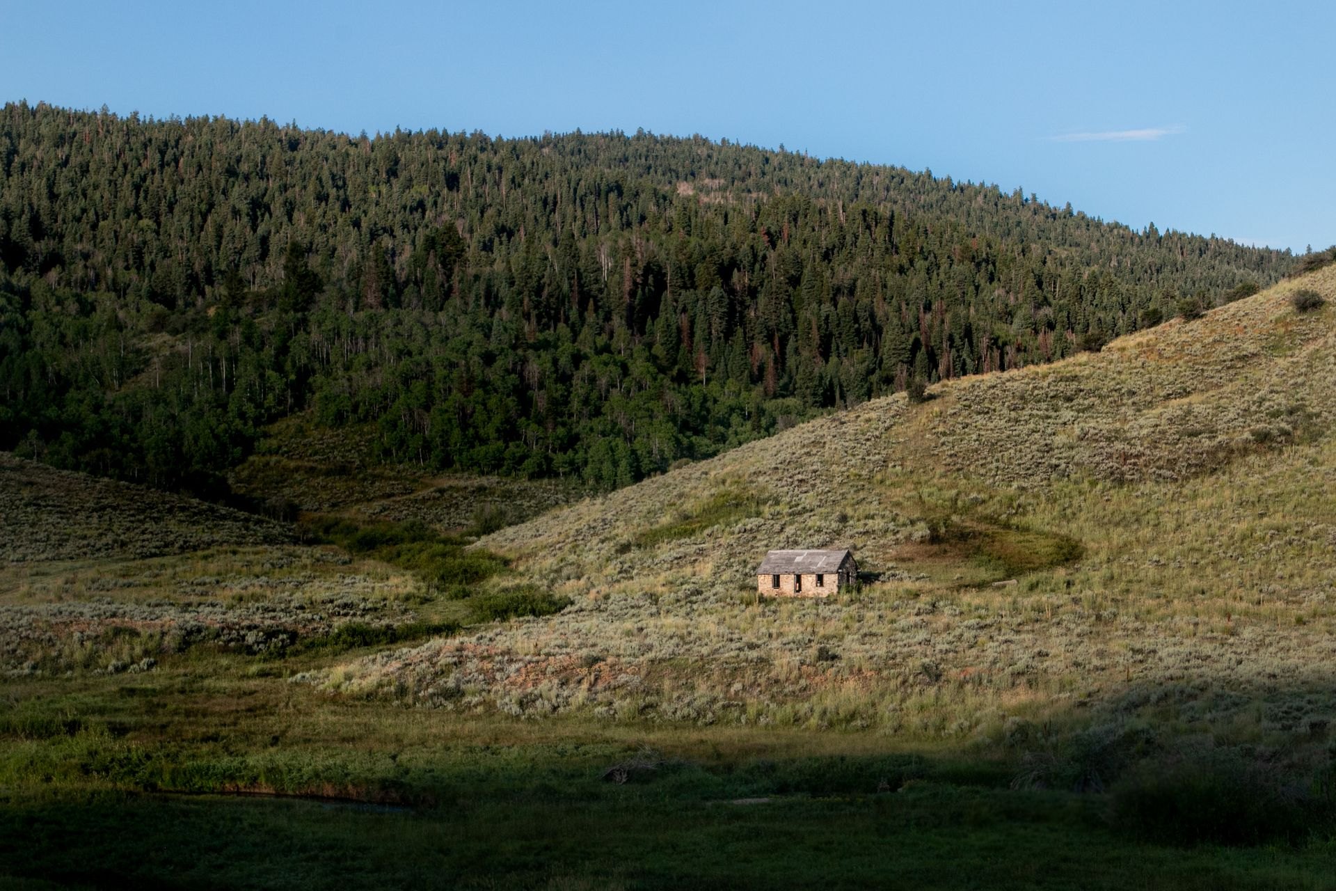 A small weathered stone cabin on a grassy hillside with forested mountains behind it.