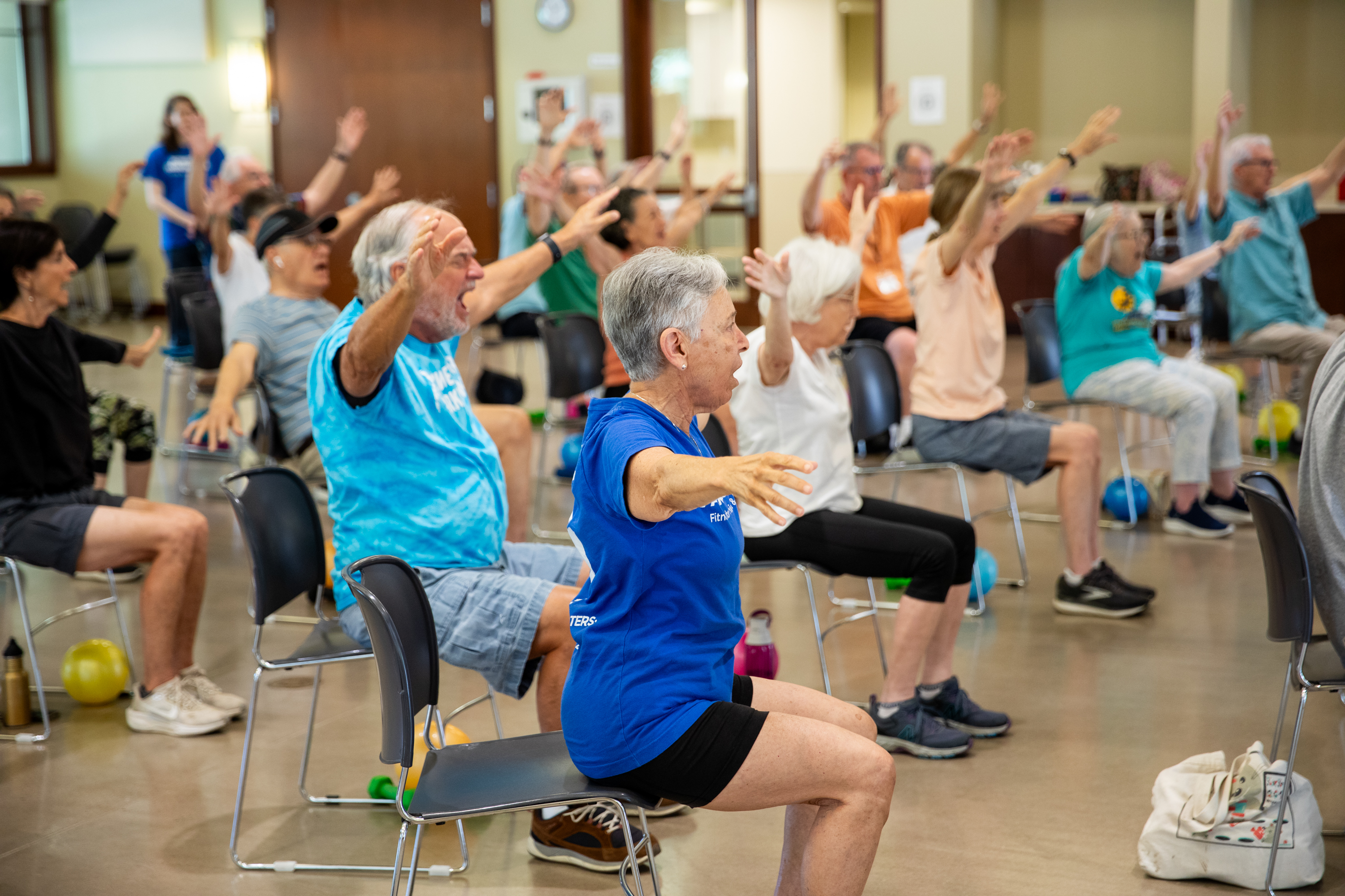 Older adults seated in chairs raise their arms during a group exercise class indoors.