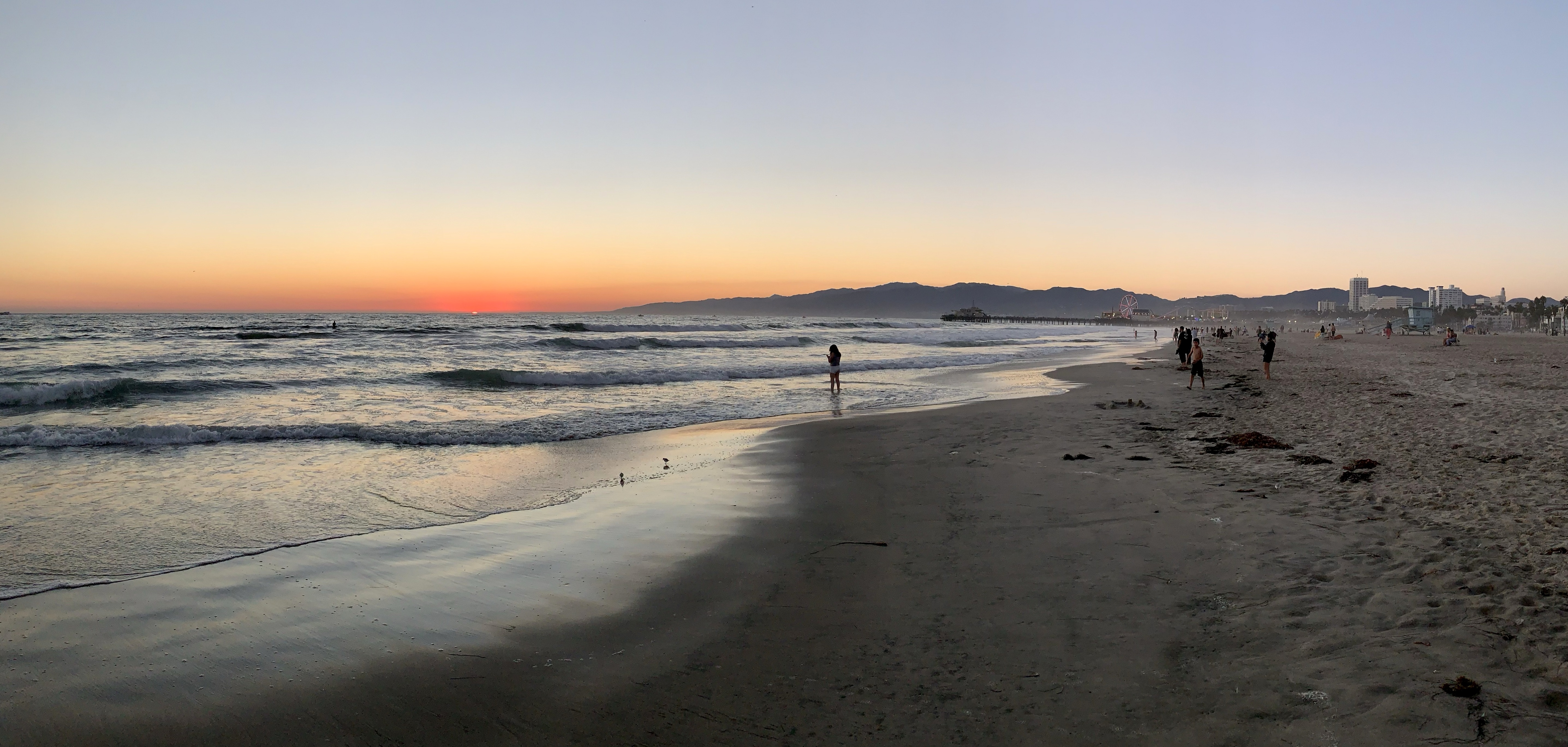 Panoramic sunset over Santa Monica beach with silhouetted beachgoers and distant mountains.