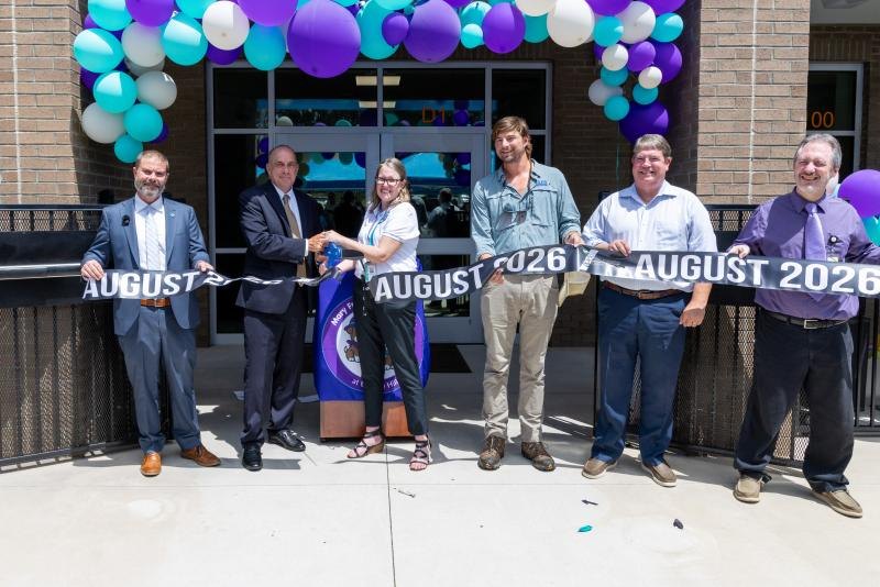Six people cut a ribbon at a building entrance decorated with teal and purple balloons.