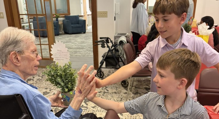 An elderly person in a wheelchair reaches out to touch hands with two young boys in a senior living common room.