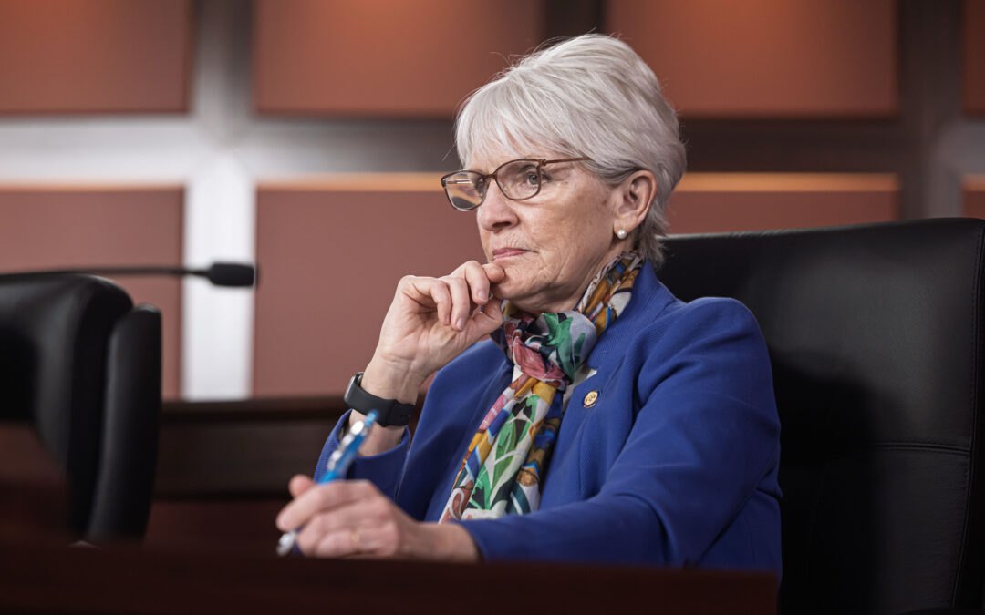 A woman with short white hair and glasses in a blue blazer seated in a legislative hearing room, holding a pen.