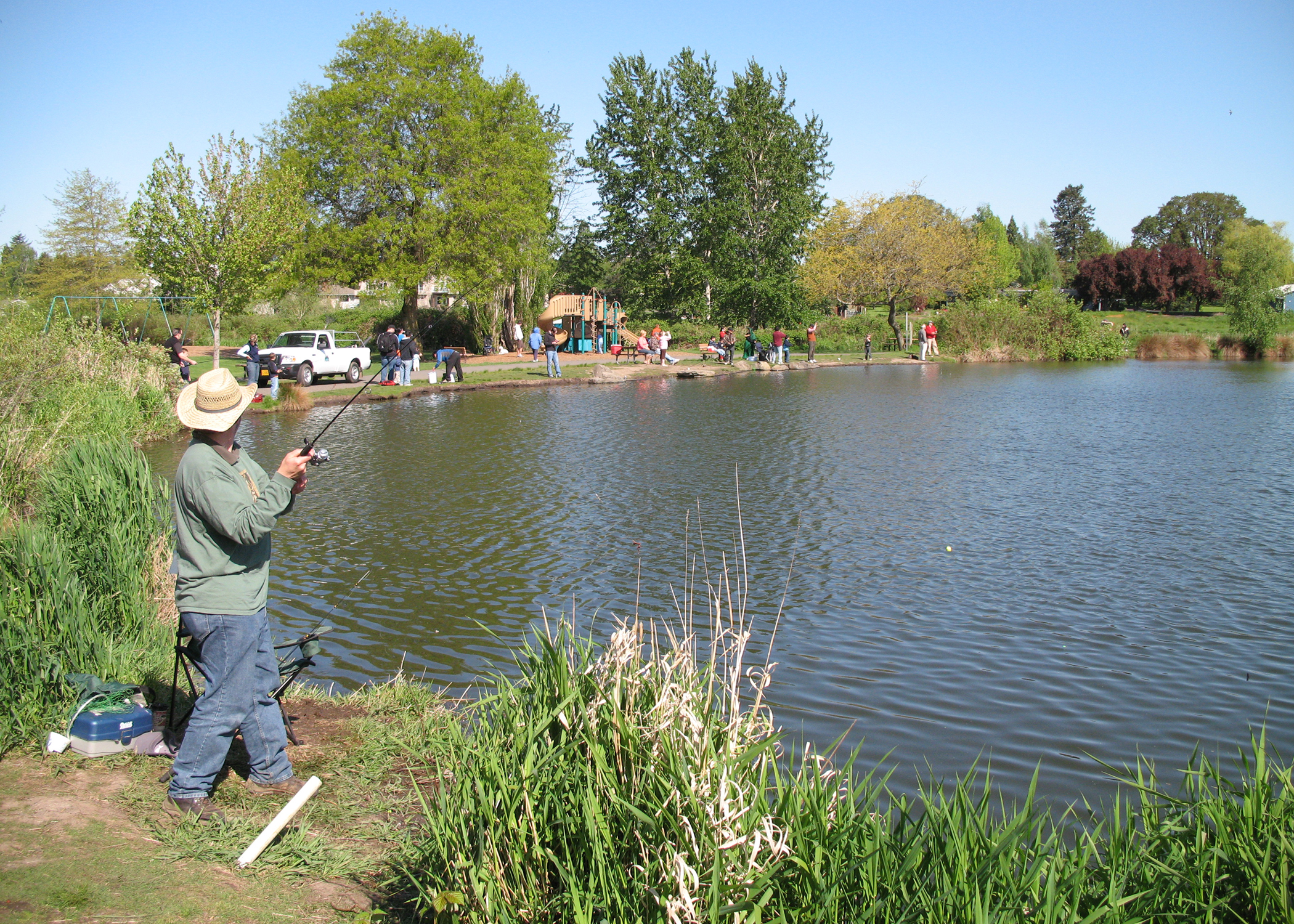 A person in a hat and jacket fishing at the edge of a calm park pond.
