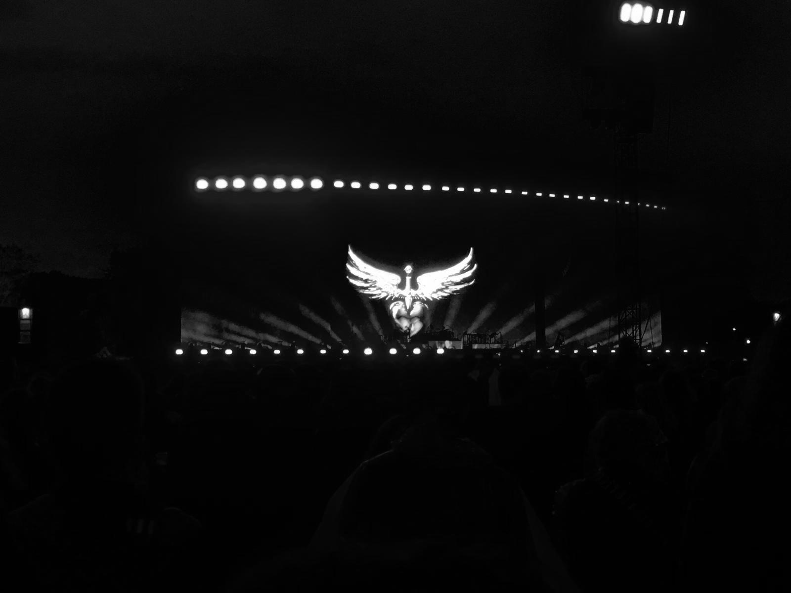 Illuminated outdoor concert stage with a large winged logo backdrop and silhouetted crowd.
