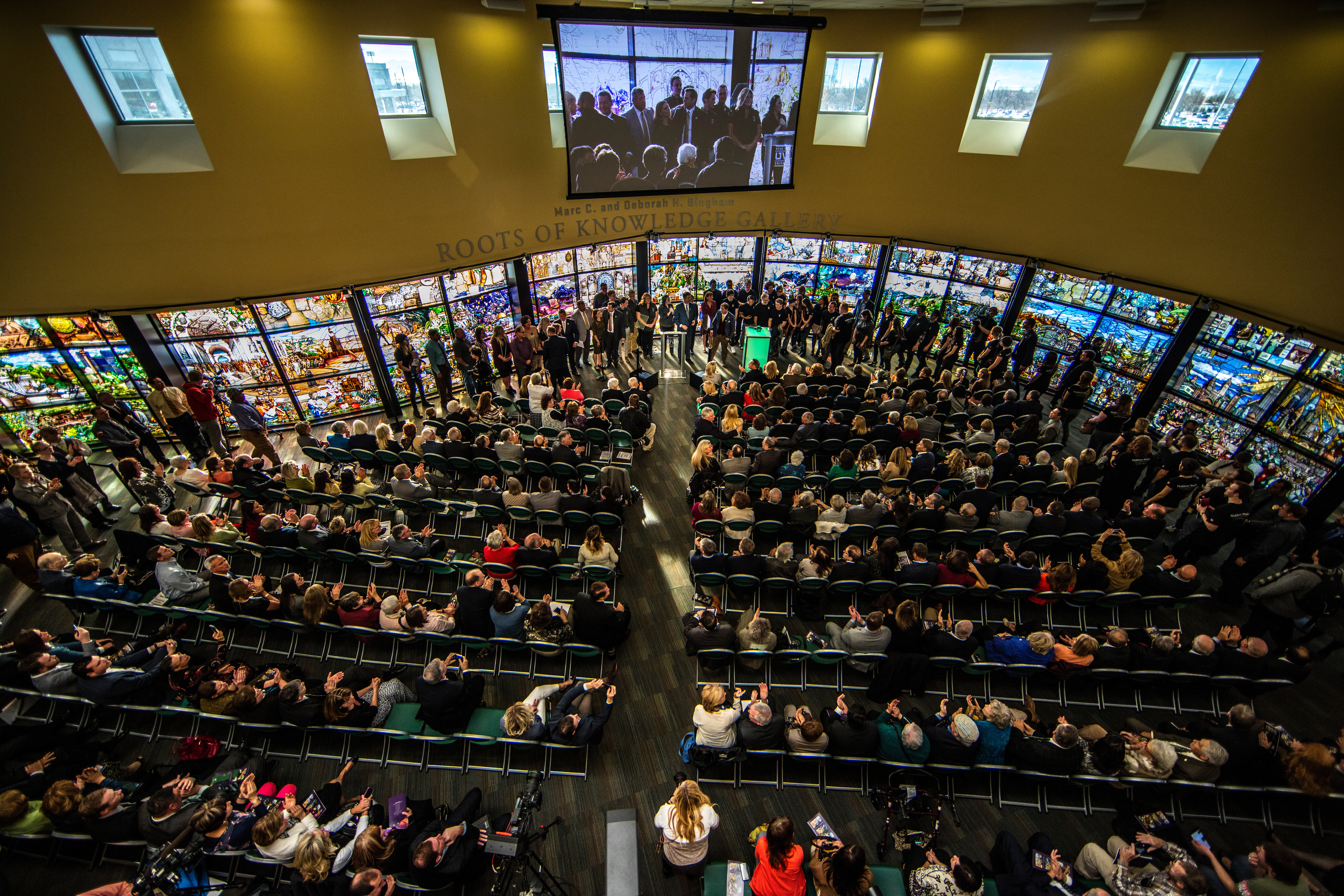 Crowd seated in a hall surrounded by tall stained-glass panels, with a large video screen above.