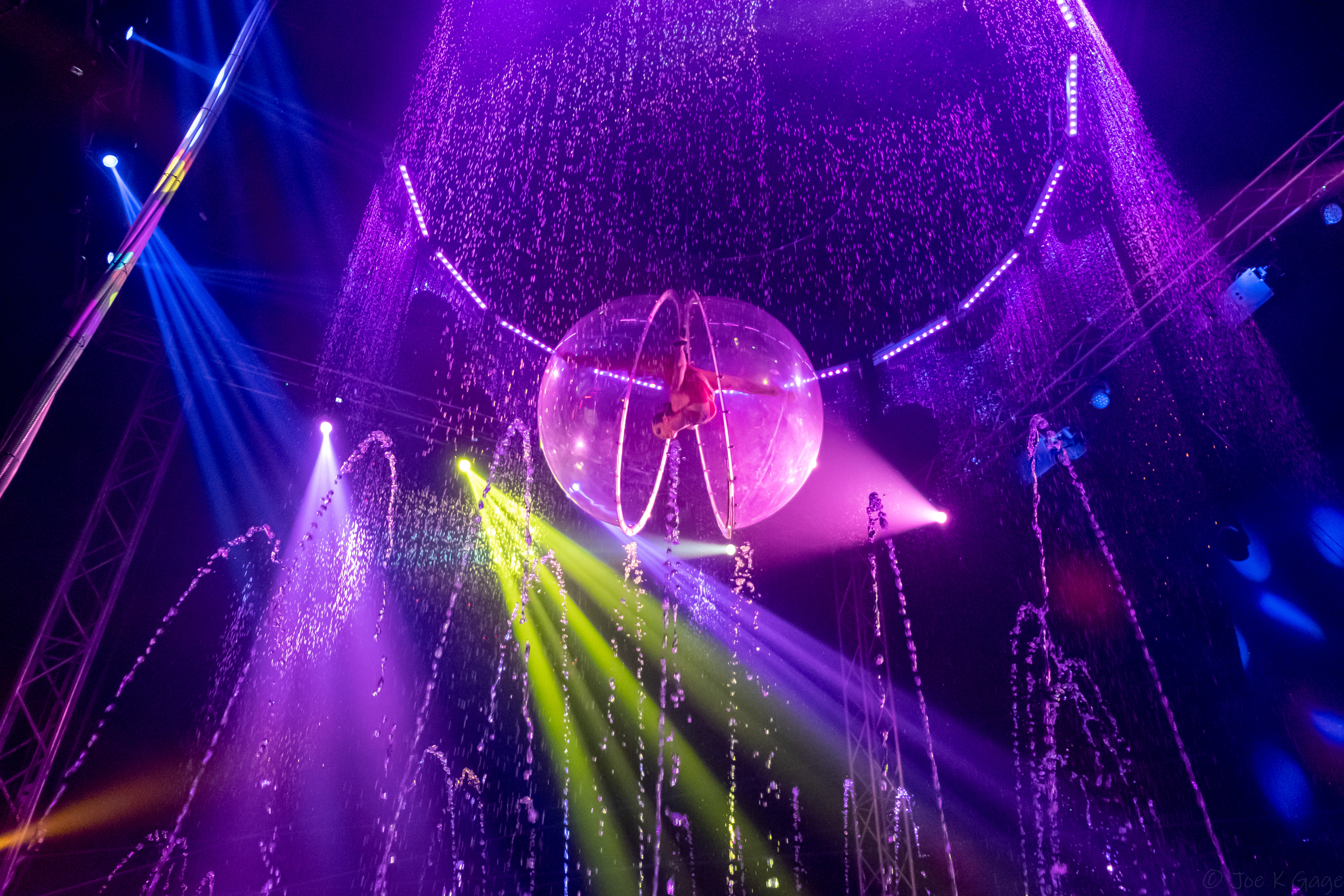 Aerialist inside a transparent sphere above a stage with water fountains and colorful lights.