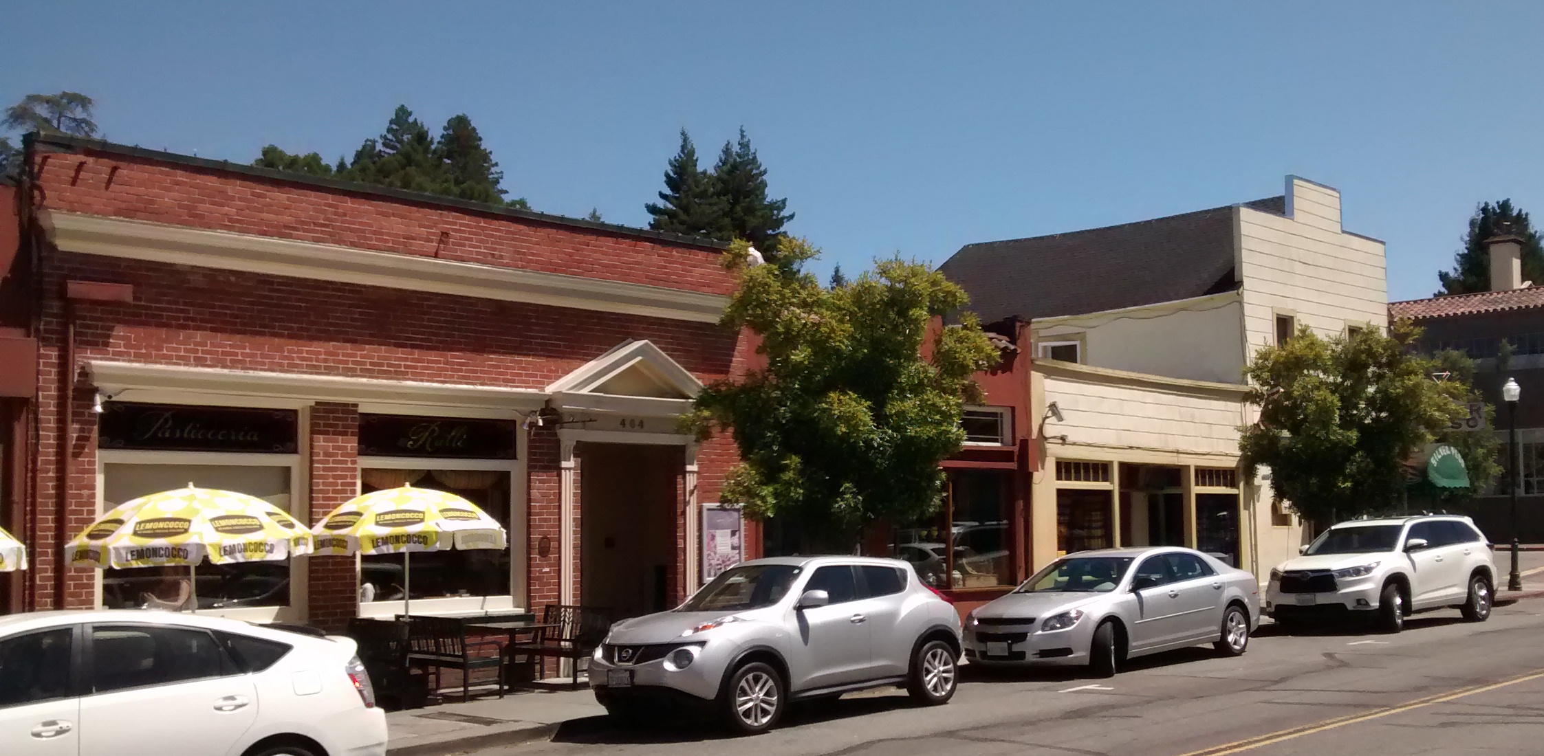 Downtown street lined with small commercial buildings, outdoor café umbrellas, and parked cars.