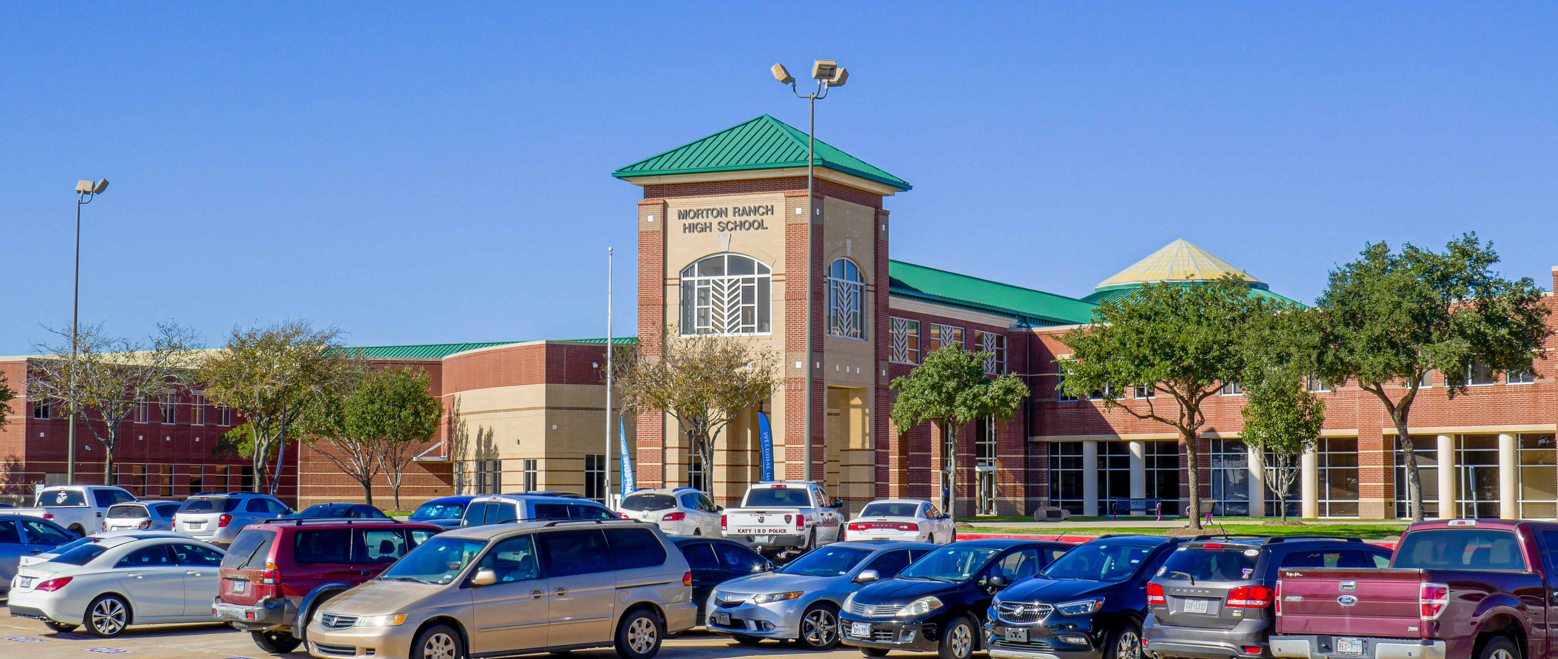 Brick exterior of a high school building with a green-roofed tower entrance and a full parking lot.