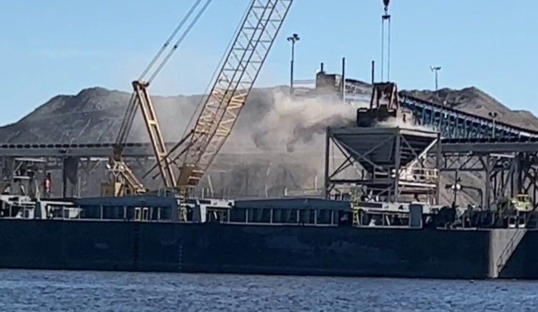 Industrial barge docked at a port terminal with cranes and dust visible in the air.