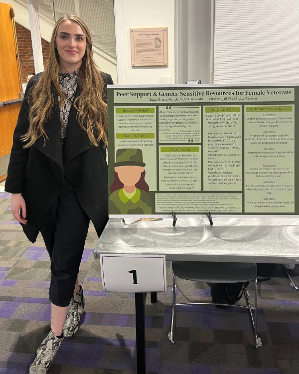Woman in a black blazer standing beside a research poster on a numbered display stand in an academic hall.