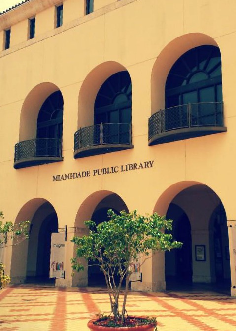 Cream-colored library building exterior with arched windows, iron balconies, and a courtyard tree.