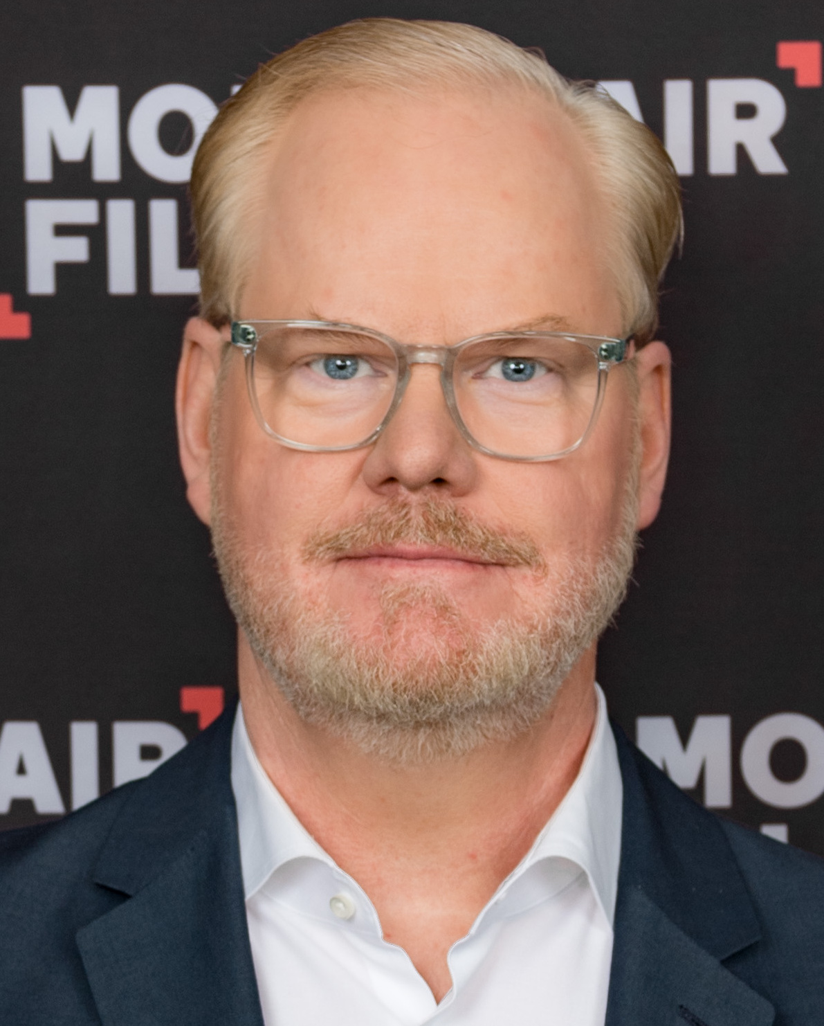 Man with blond hair and glasses in a navy blazer stands before an event backdrop.