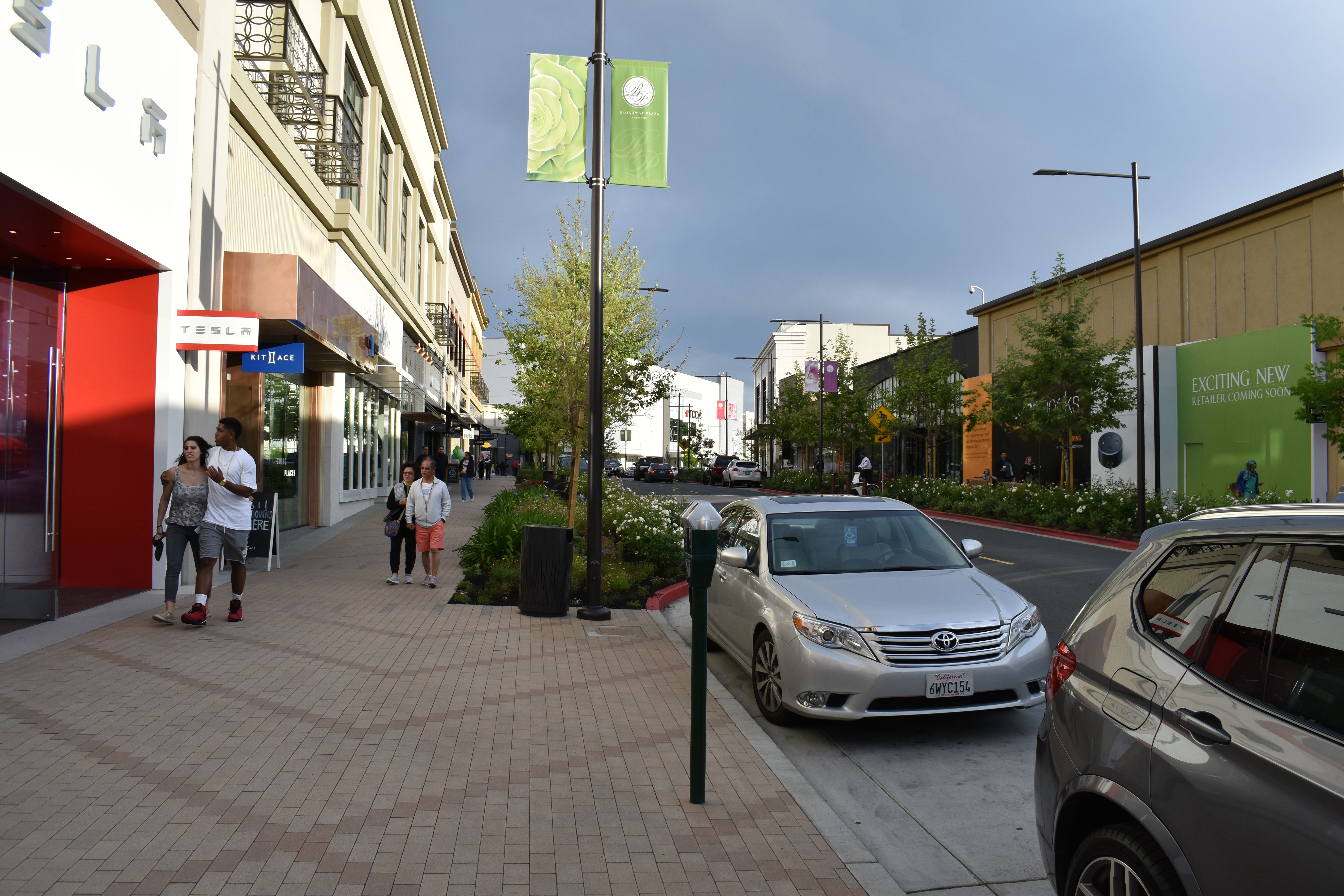 Pedestrian walkway lined with retail storefronts and green banners at an outdoor shopping center.