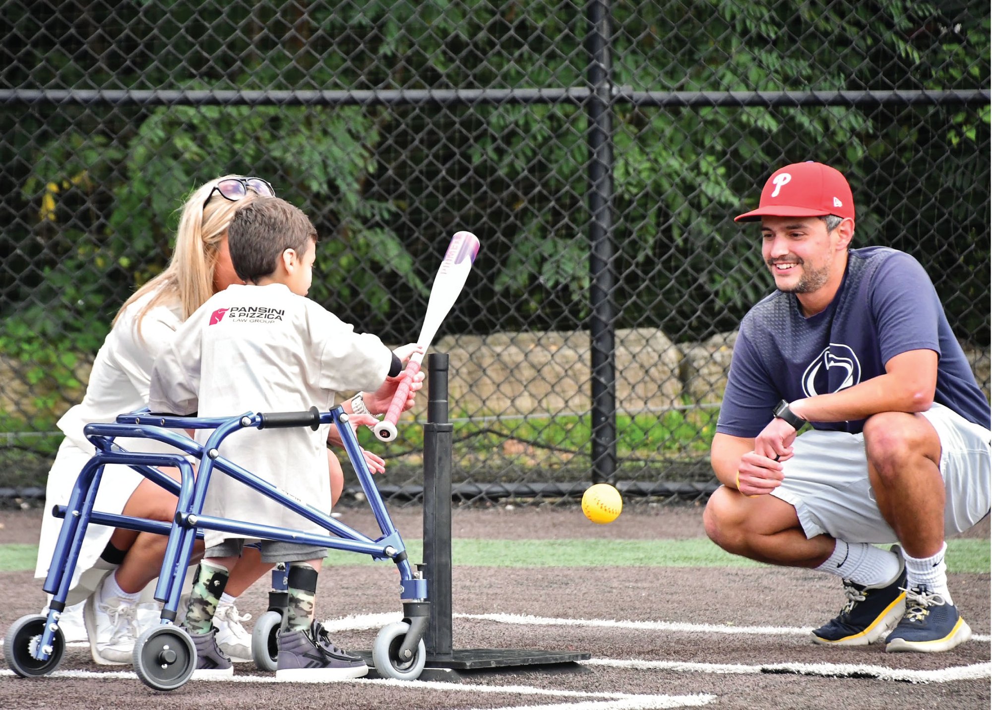 Child using a walker swings a bat at a tee while an adult crouches nearby on a baseball field.