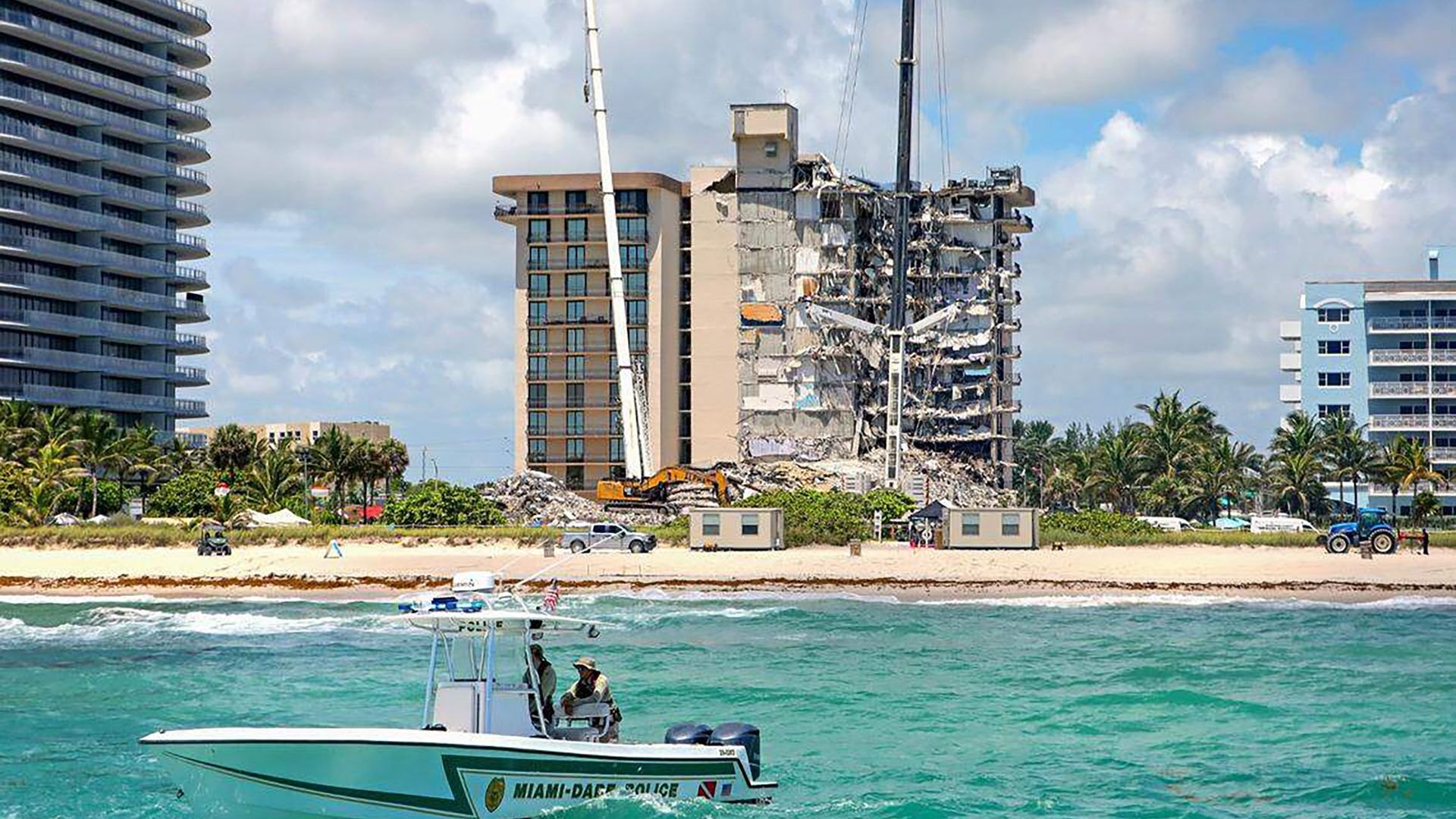 Partially collapsed high-rise condominium building viewed from the ocean with a police boat and crane visible.