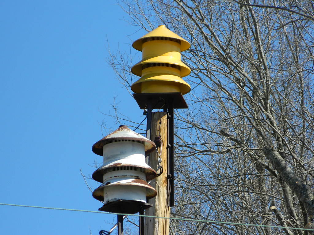 Two outdoor warning sirens mounted on a wooden utility pole against a blue sky.