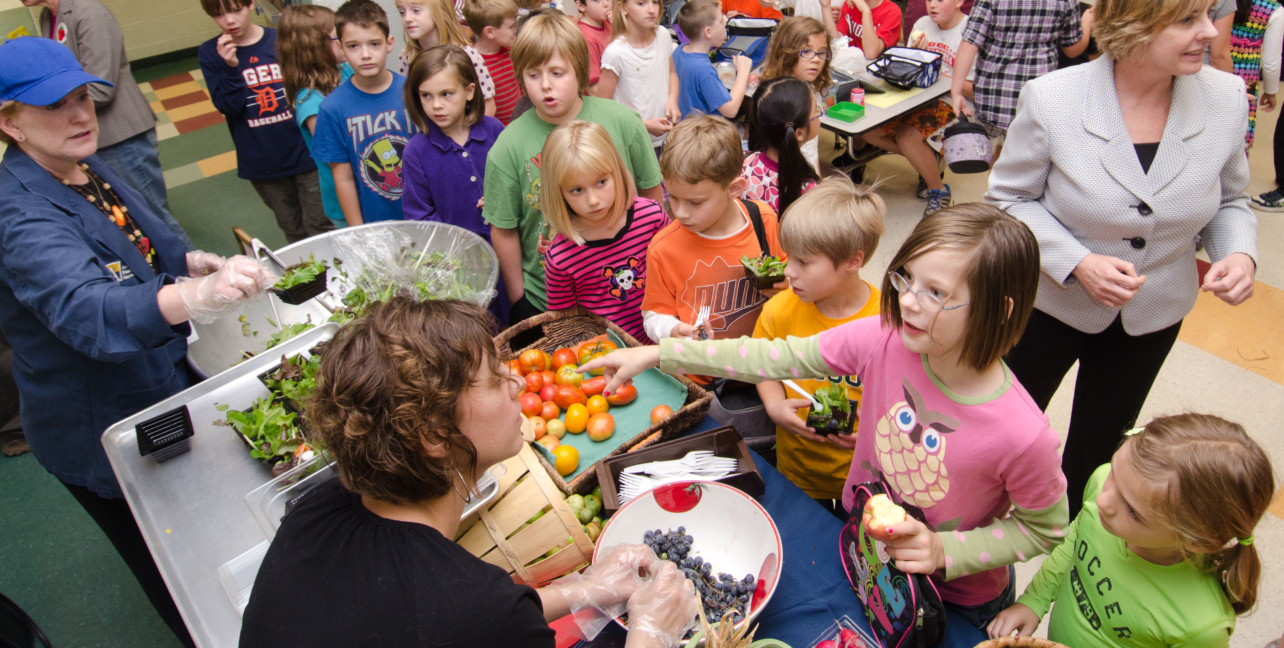 Children gathered around a table with baskets of fresh greens, tomatoes, and vegetables.
