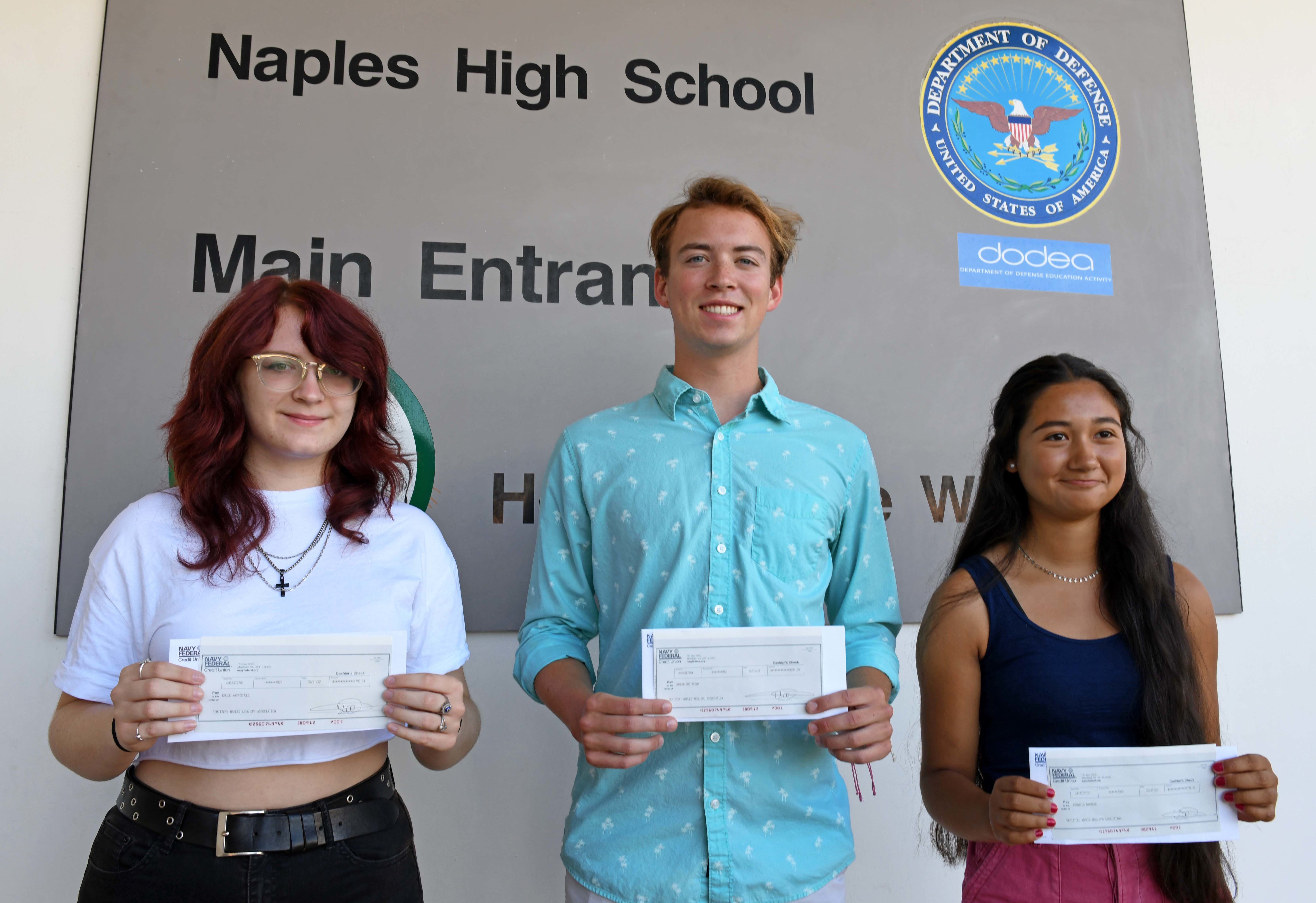 Three teenagers holding scholarship checks in front of a school entrance sign.
