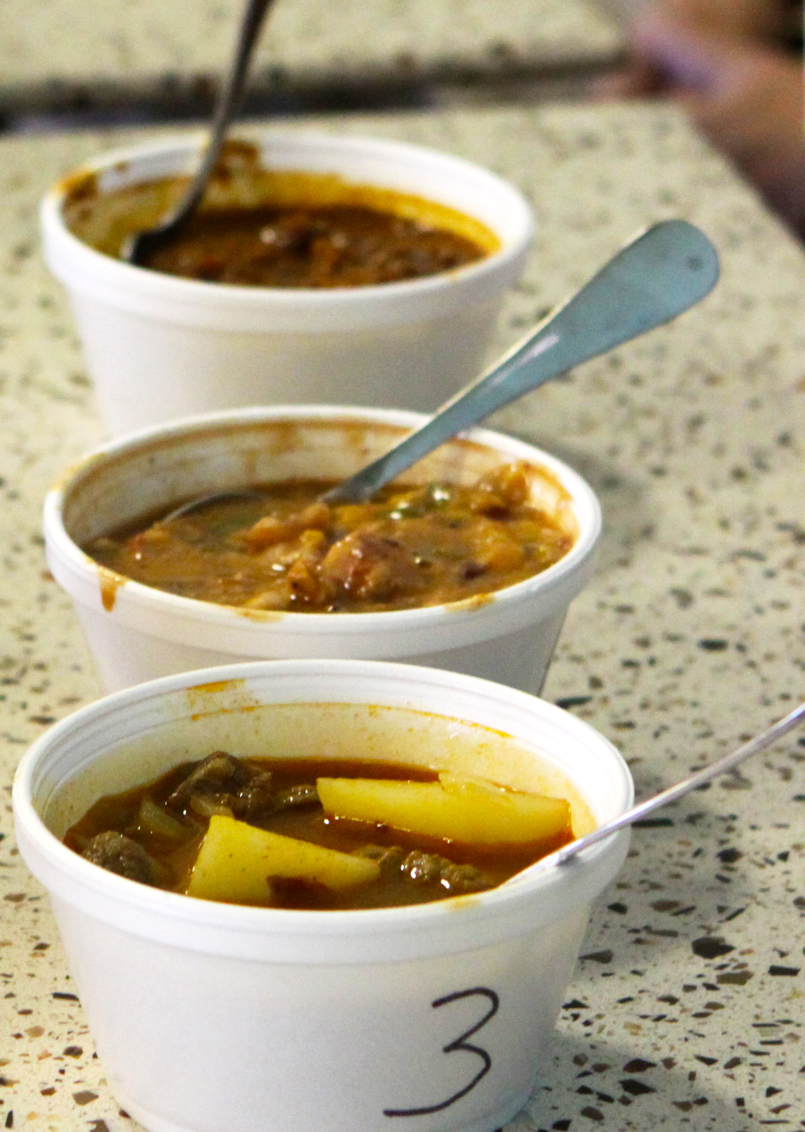 Three numbered styrofoam cups of chili with spoons lined up on a countertop.