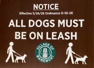 Brown sign reading 'ALL DOGS MUST BE ON LEASH' with village seal and dog-walking pictograms.