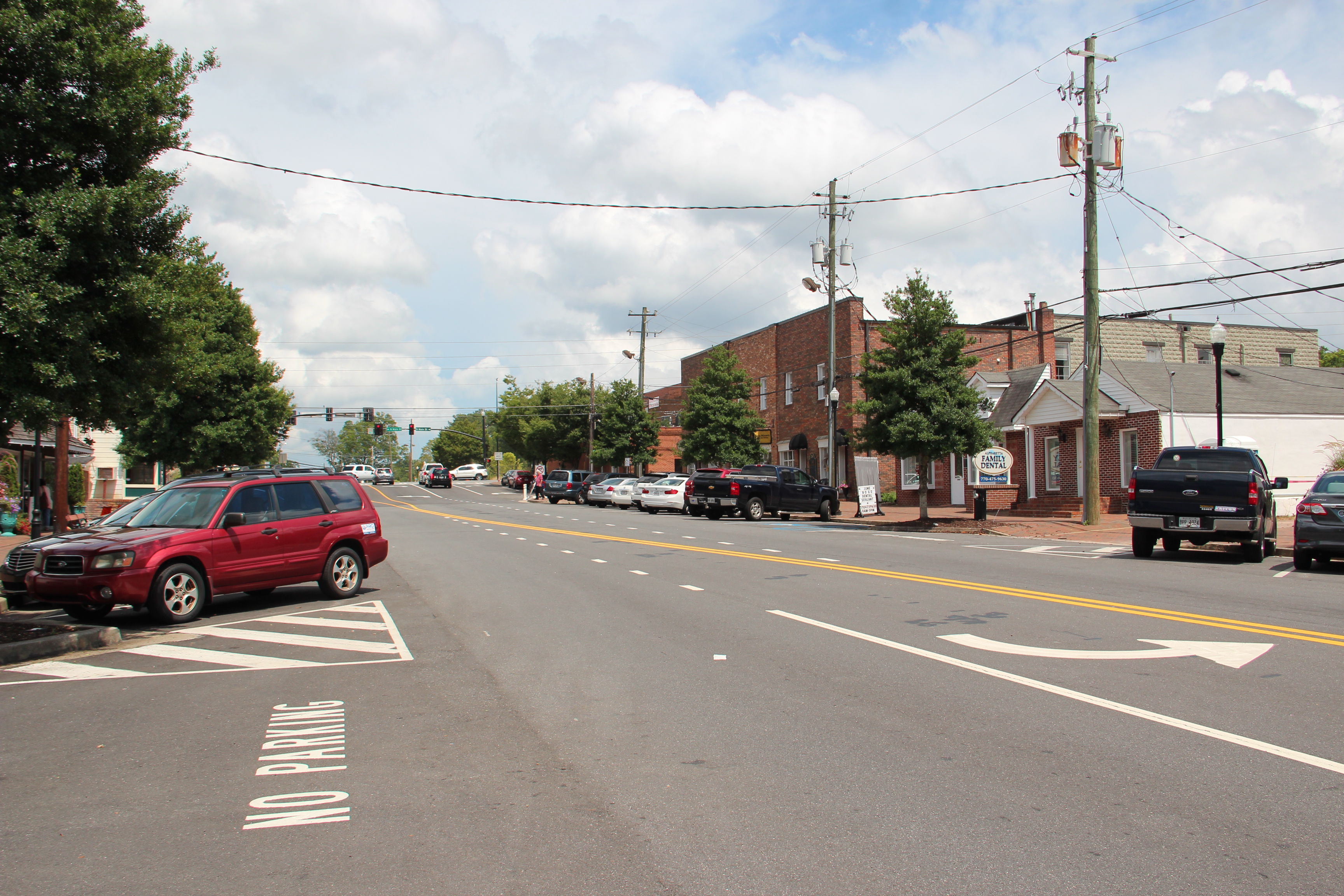 A two-lane main street in a small downtown with brick buildings, parked cars, and trees.