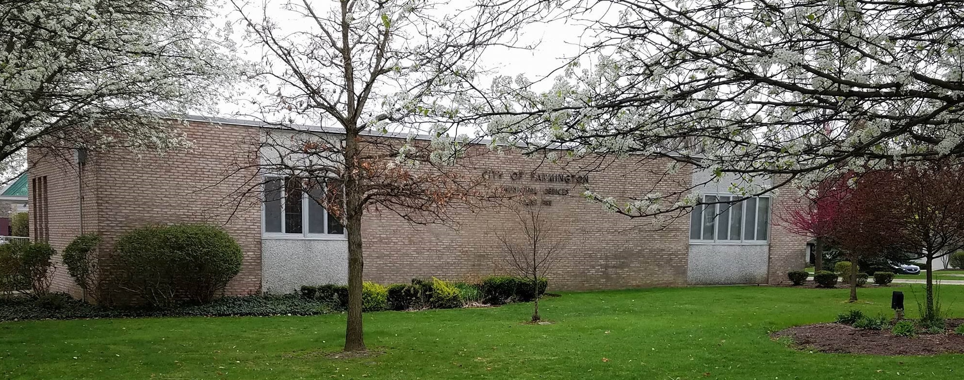 Single-story brick municipal building behind blooming spring trees and a green lawn.