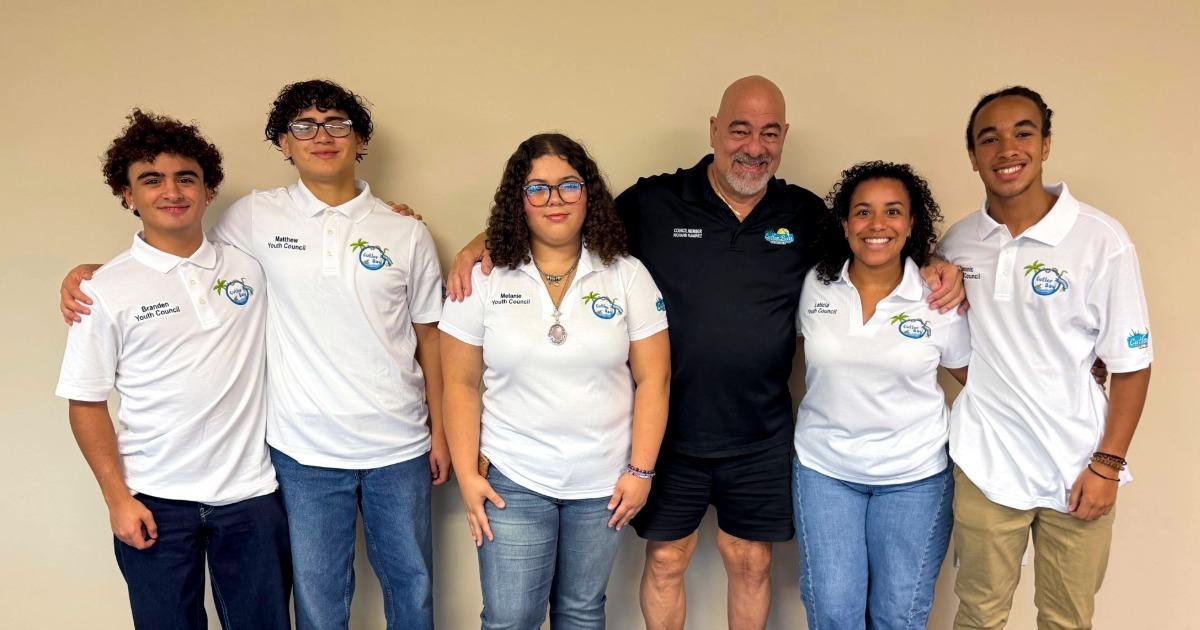 Six people in matching white polo shirts pose together indoors against a beige wall.