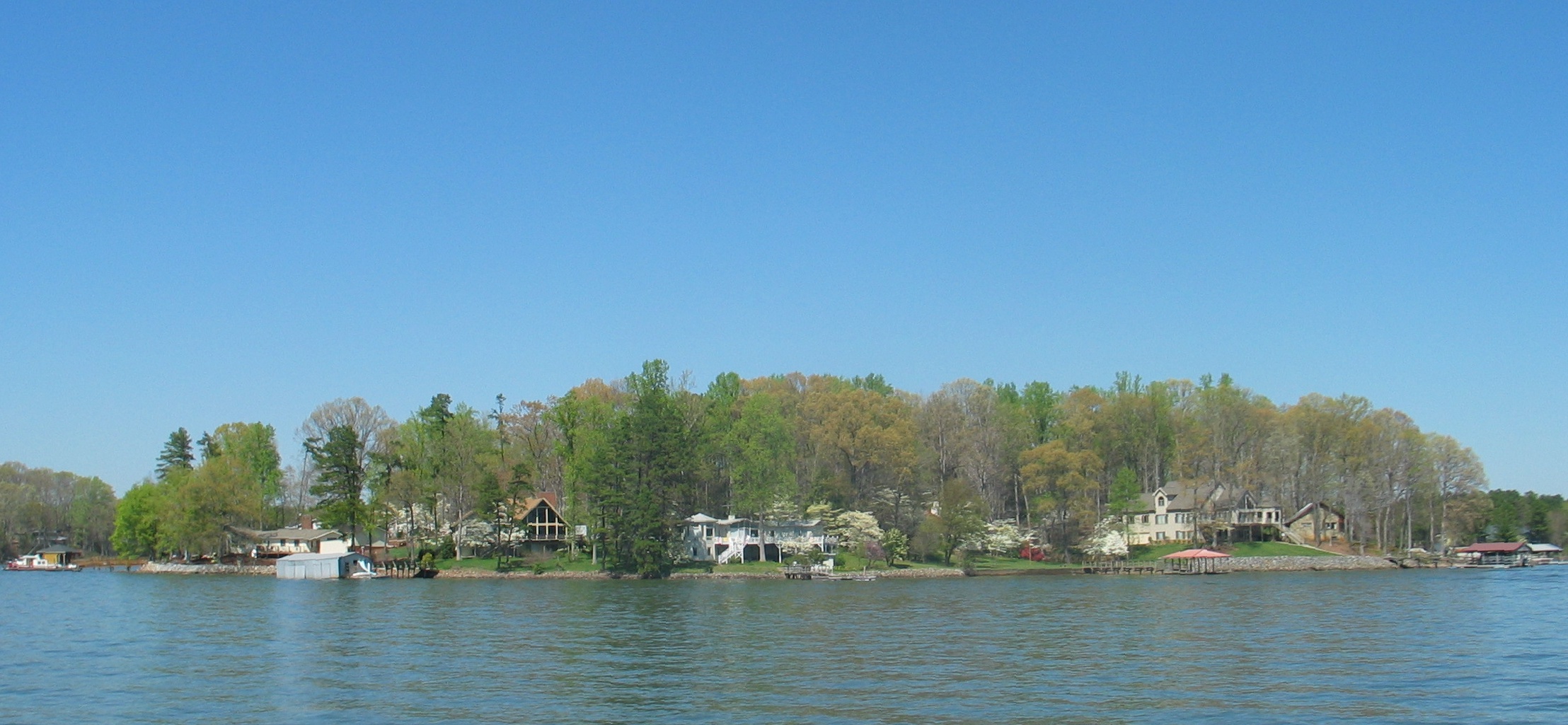 Calm lake shoreline with lakefront homes and wooden docks among leafy trees.
