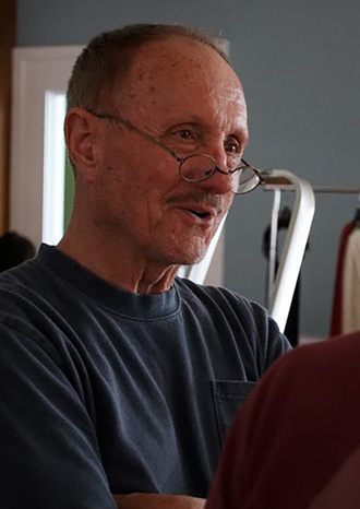 Older man with gray hair and wire-rimmed glasses, wearing a blue t-shirt, speaking indoors.