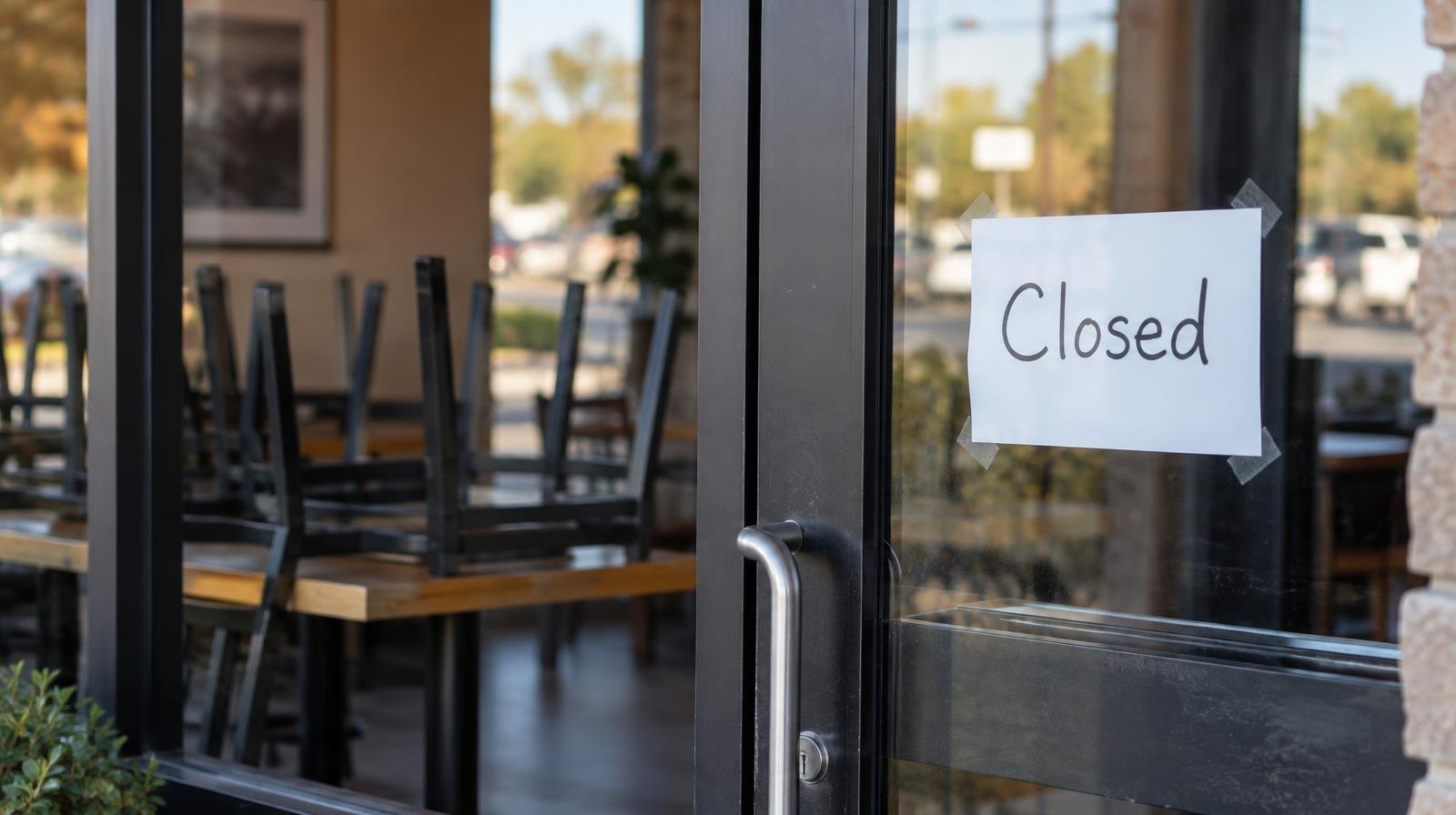 Glass restaurant door with a closed sign, chairs stacked on tables inside.