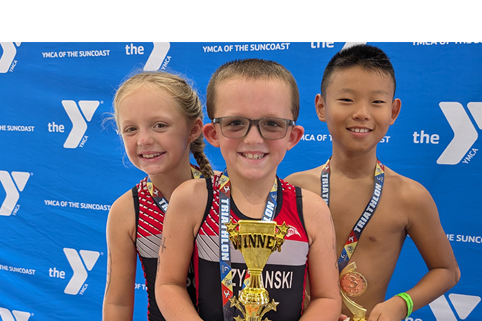 Three children in athletic gear pose with a trophy and medals in front of a YMCA backdrop.