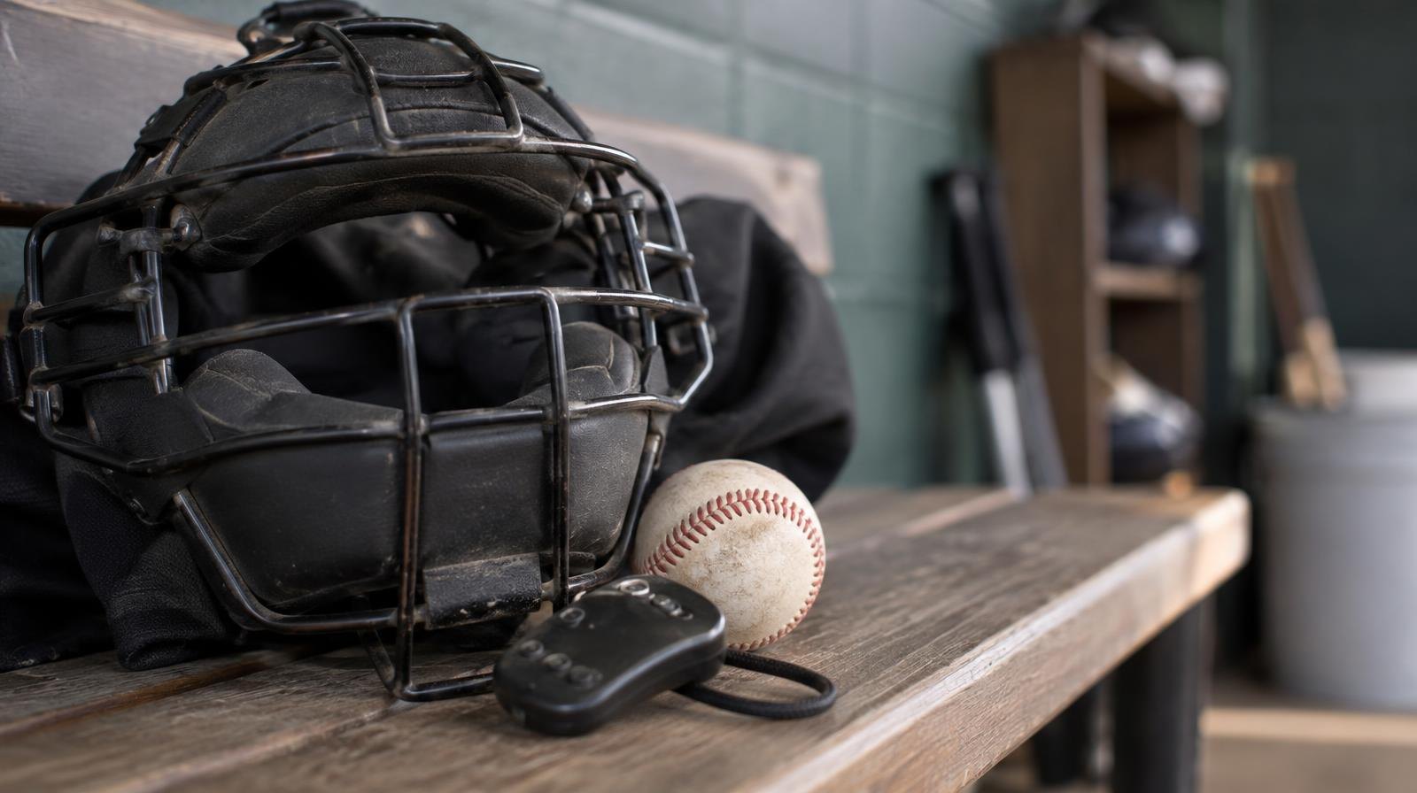 A baseball umpire's mask and pitch indicator clicker rest on a wooden bench beside a baseball.