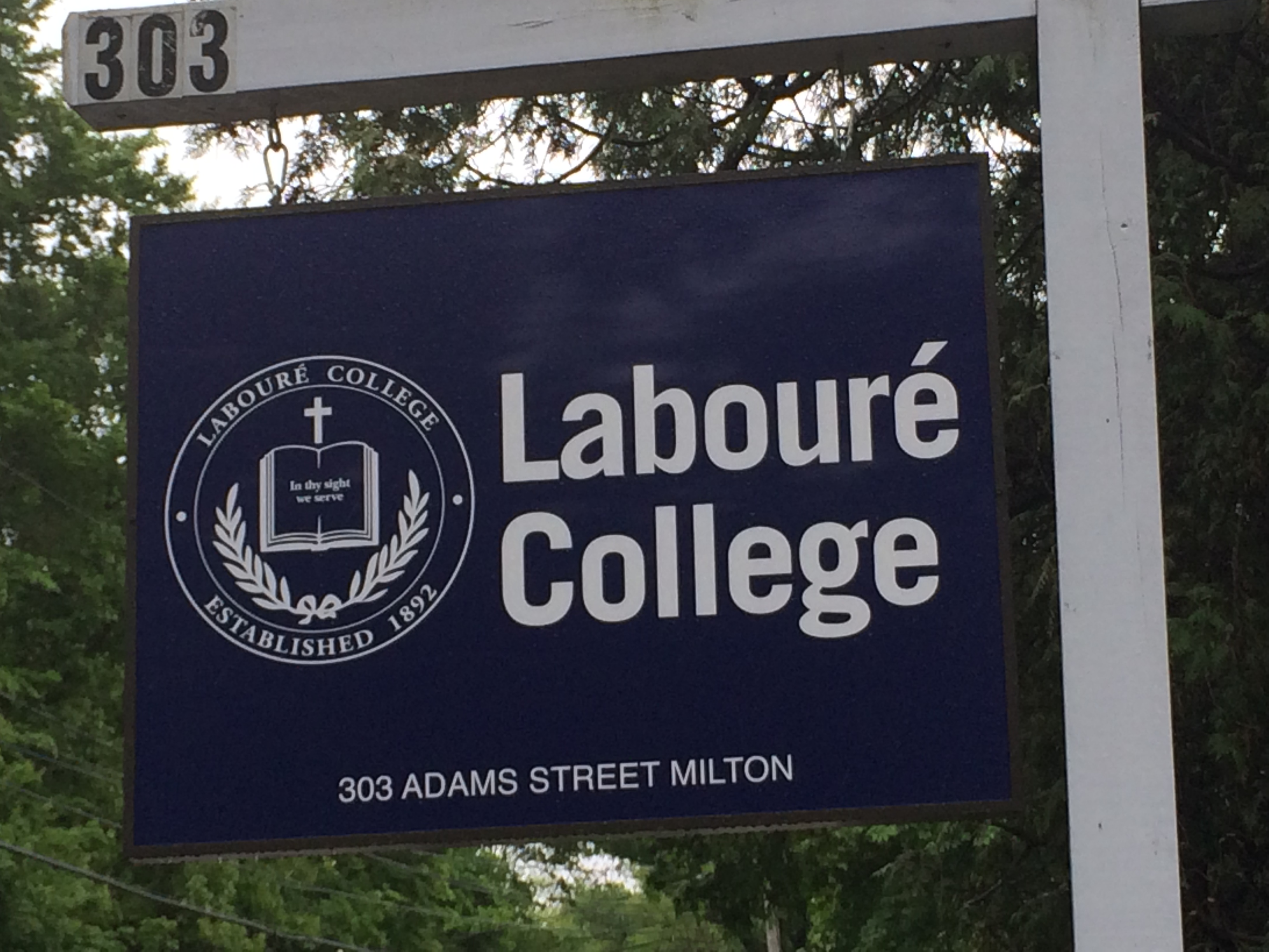 Navy blue college sign on a white post reading 303 Adams Street, Milton, surrounded by trees.