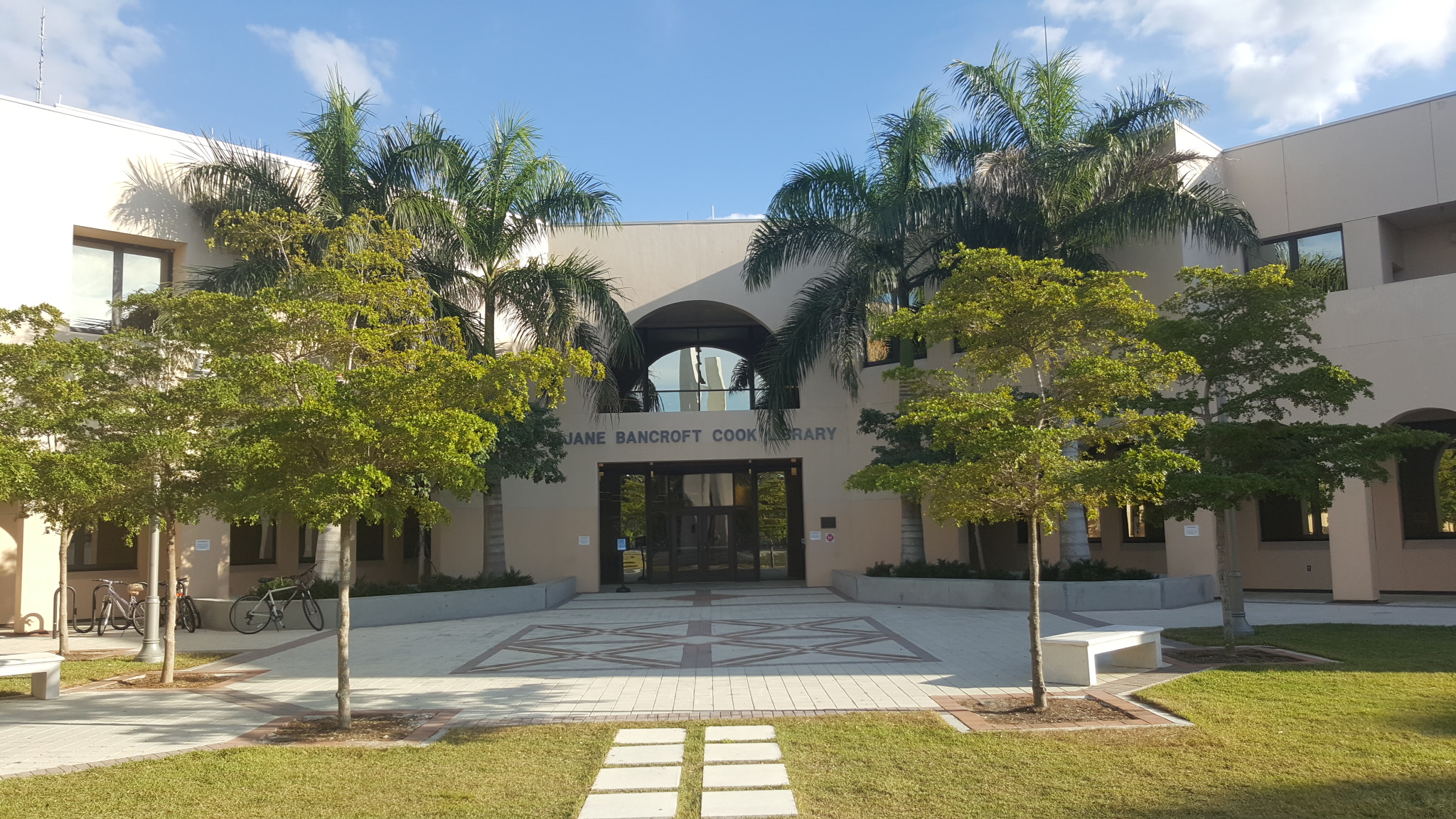 Library building exterior with arched entrance, palm trees, and a decorative tiled plaza.