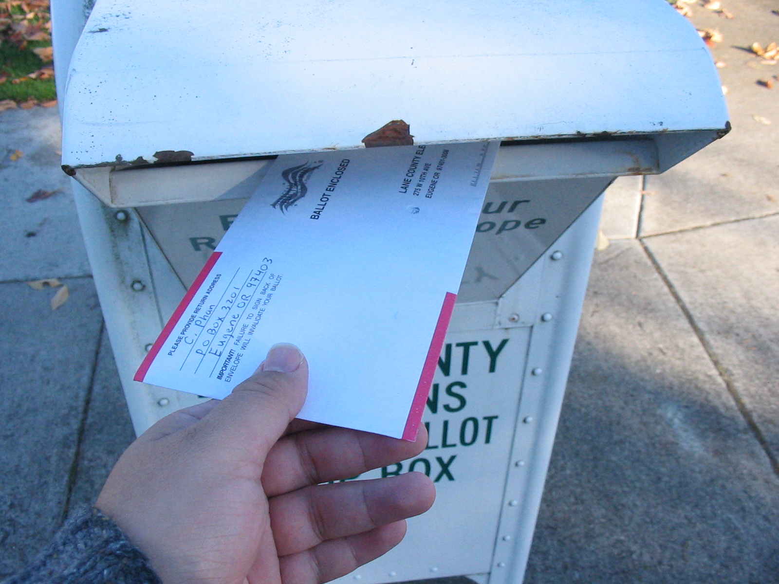 A hand inserting a mail ballot envelope into a white outdoor county ballot drop box.