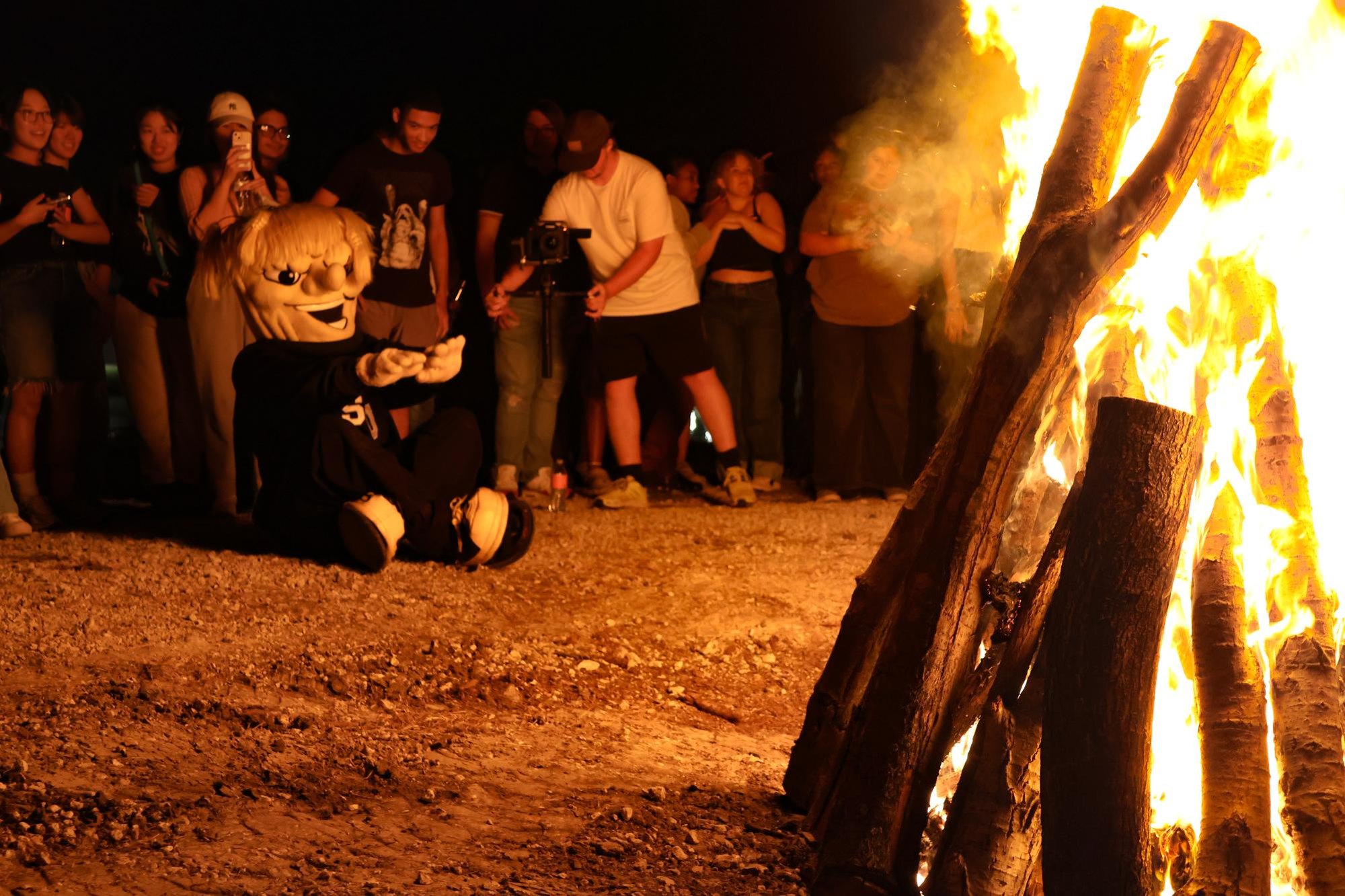 A large outdoor bonfire burns at night with a crowd of spectators gathered behind it.
